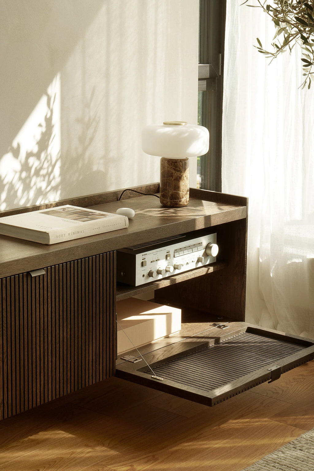 TAMO Meda 2 TV Unit, displayed in a sunlit living room, featuring rich dark brown wood with ribbed panels, a pull-down slatted door, and an open shelf holding a vintage stereo. A premium minimalist design for modern home entertainment.