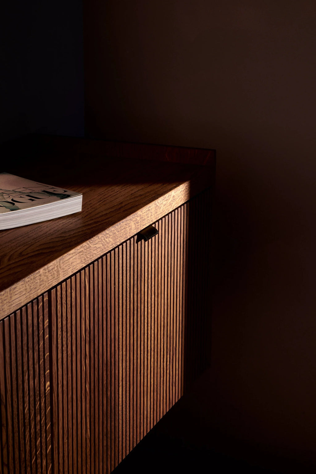 TAMO Meda 2 TV Unit, a dark rich brown oak wood cabinet with fluted paneling and a black leather handle, styled with a magazine on top in a moody, low-lit setting. Perfect for a sophisticated living room.