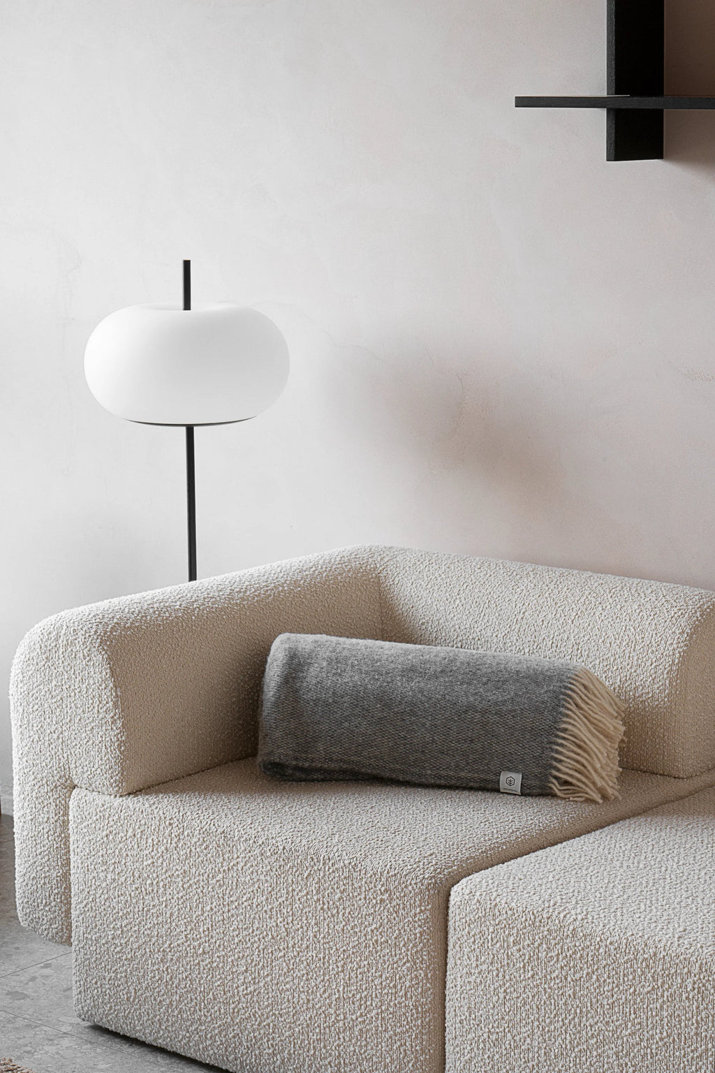 TAMO Meri Blanket Throw: A luxurious rolled gray melange wool throw with light beige fringe, draped on a textured off-white sofa in a lifestyle setting. Perfect for cozy home decor.