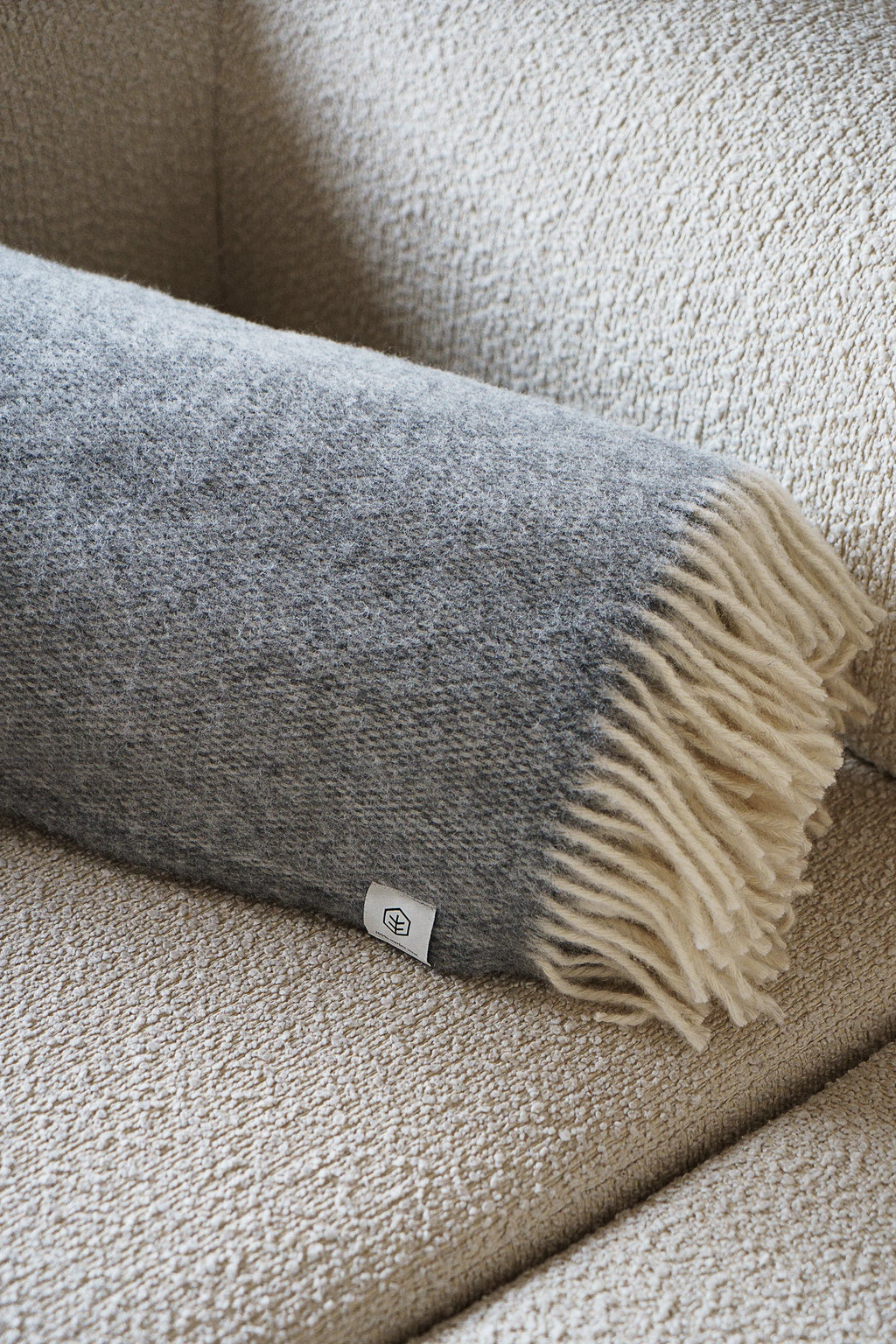 TAMO Meri Blanket Throw, a premium grey marl wool throw with natural off-white fringes, partially rolled on a neutral textured couch. A cozy home accessory.