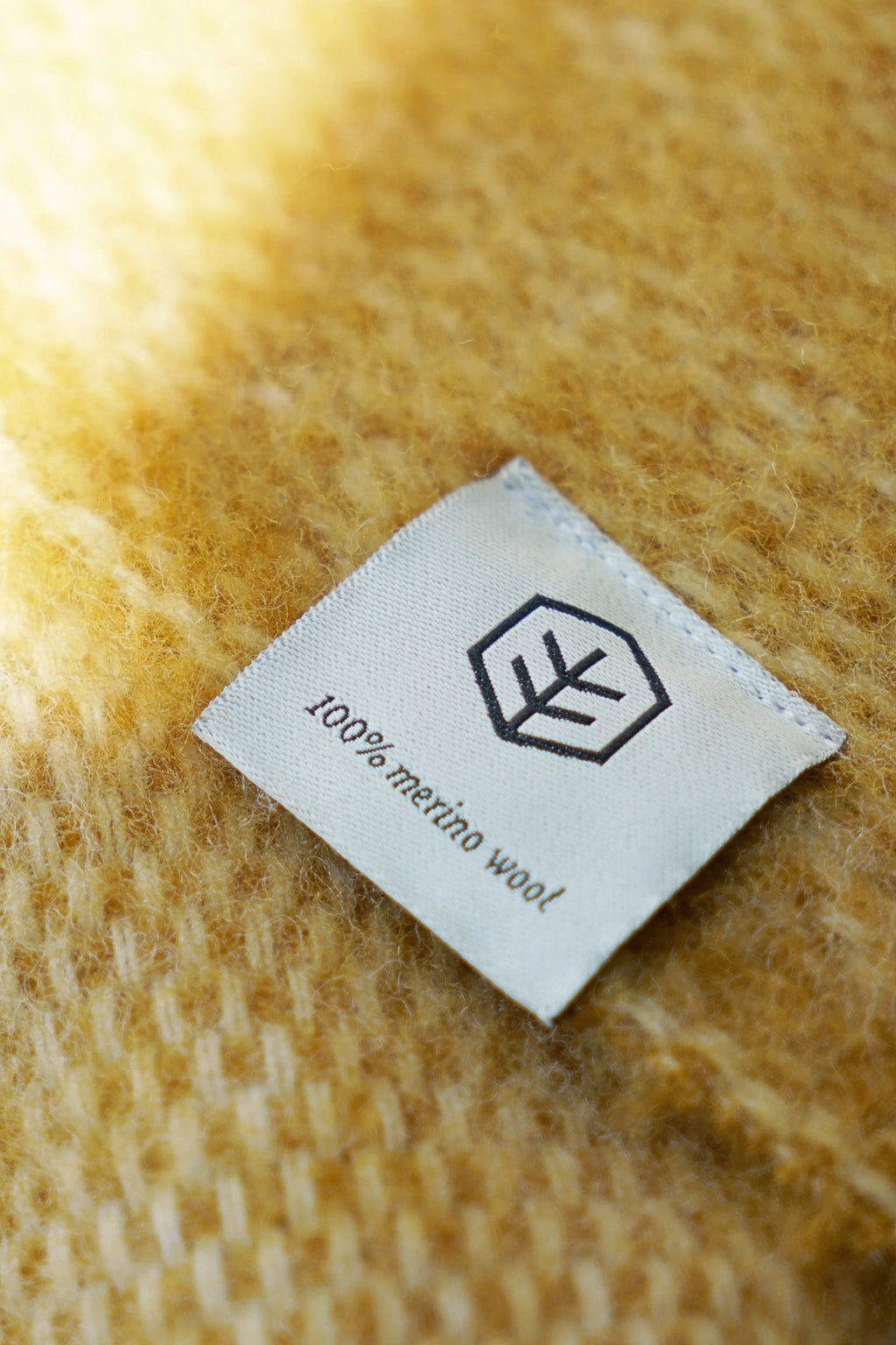 TAMO Meri Blanket Throw: Close-up of a cream-colored woven label on a premium amber yellow 100% merino wool blanket, showing visible soft, fluffy texture. Perfect for luxurious comfort.