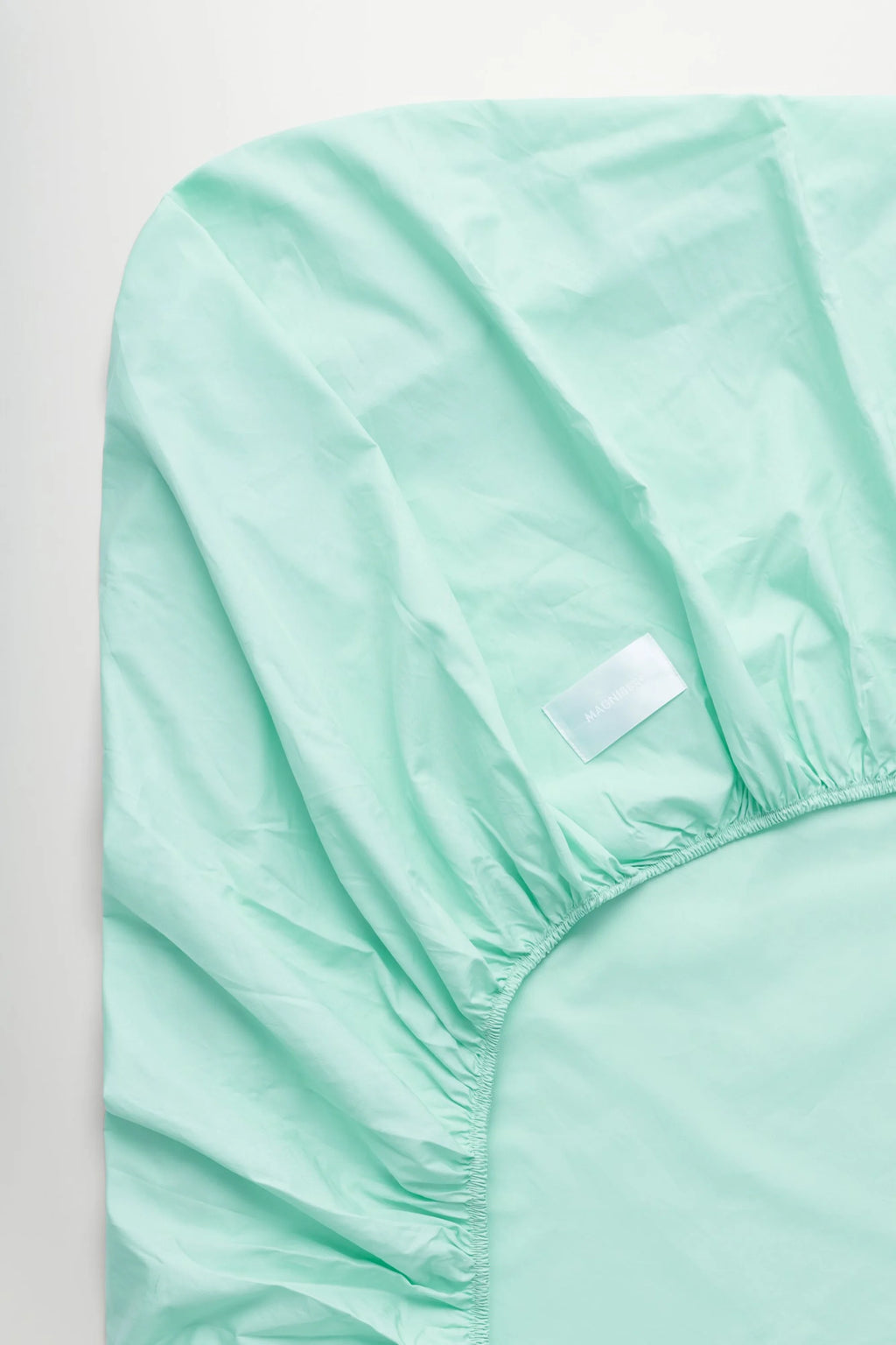 Magniberg Mother Poplin Fitted Sheet, a pastel aqua fitted sheet crafted from crisp cotton poplin fabric, showcasing its smooth texture and gently rumpled surface with a visible elasticized corner. This minimalist bedding is ideal for creating a serene and contemporary look in a modern bedroom or Scandinavian-style interior.