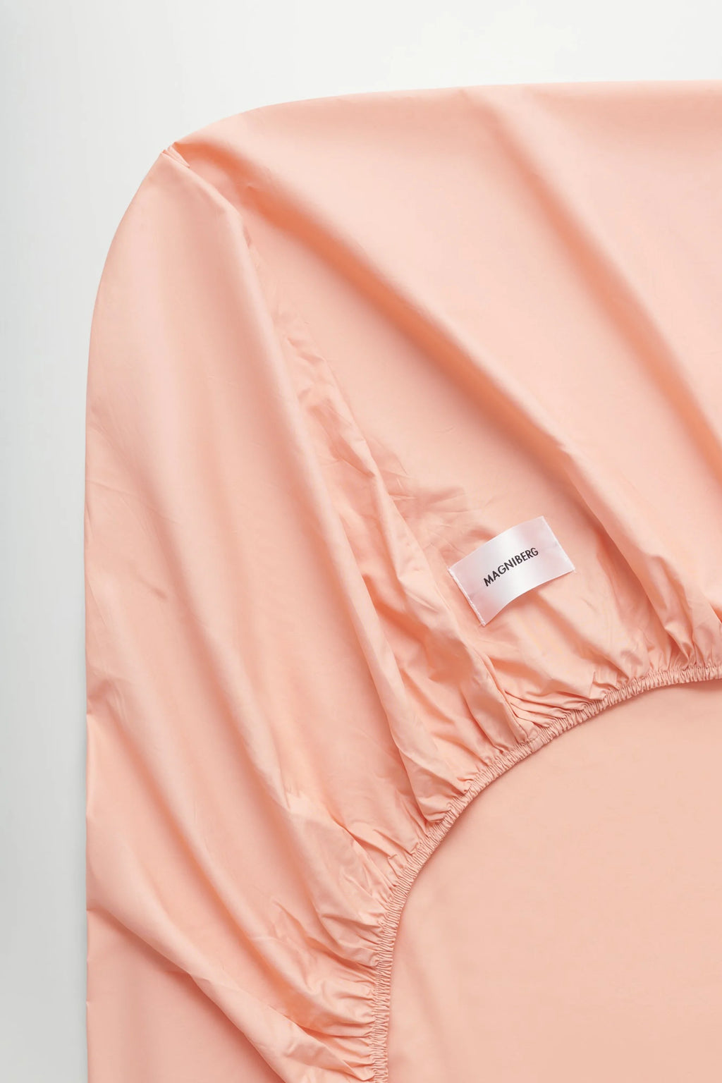 Magniberg Mother Poplin Fitted Sheet, a luxurious soft peach cotton poplin fitted sheet with a crisp, smooth finish and fully elasticized edges for a secure fit. This modern minimalist bedding essential is perfect for creating a serene and contemporary bedroom aesthetic.