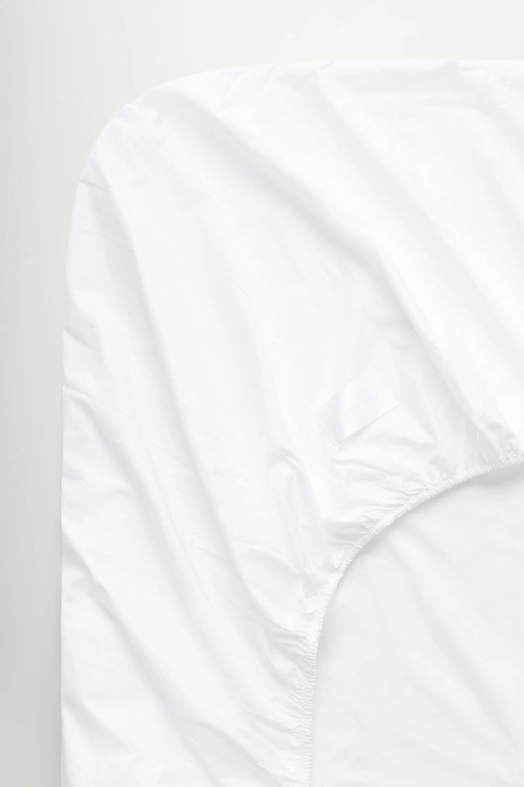 Magniberg Mother Poplin Fitted Sheet, a crisp white fitted sheet crafted from smooth cotton poplin fabric, showcasing subtle natural folds and a tightly elasticized edge for a secure mattress fit. This minimalist and classic white bedding is ideal for creating a serene bedroom sanctuary.