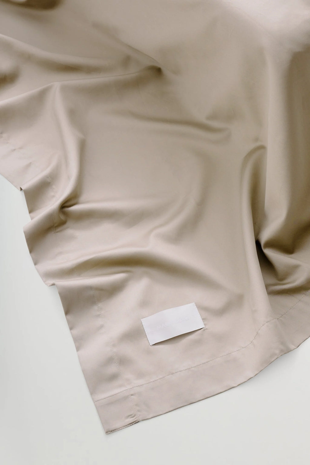Magniberg Mother Sateen Flat Sheet, a luxurious warm beige sateen flat sheet crafted from premium cotton with a smooth, soft texture and subtle sheen, showcasing elegant draping and meticulous stitching along the hem. This contemporary bedding piece is ideal for creating a modern minimalist bedroom aesthetic or a sophisticated, neutral-toned sleep sanctuary.