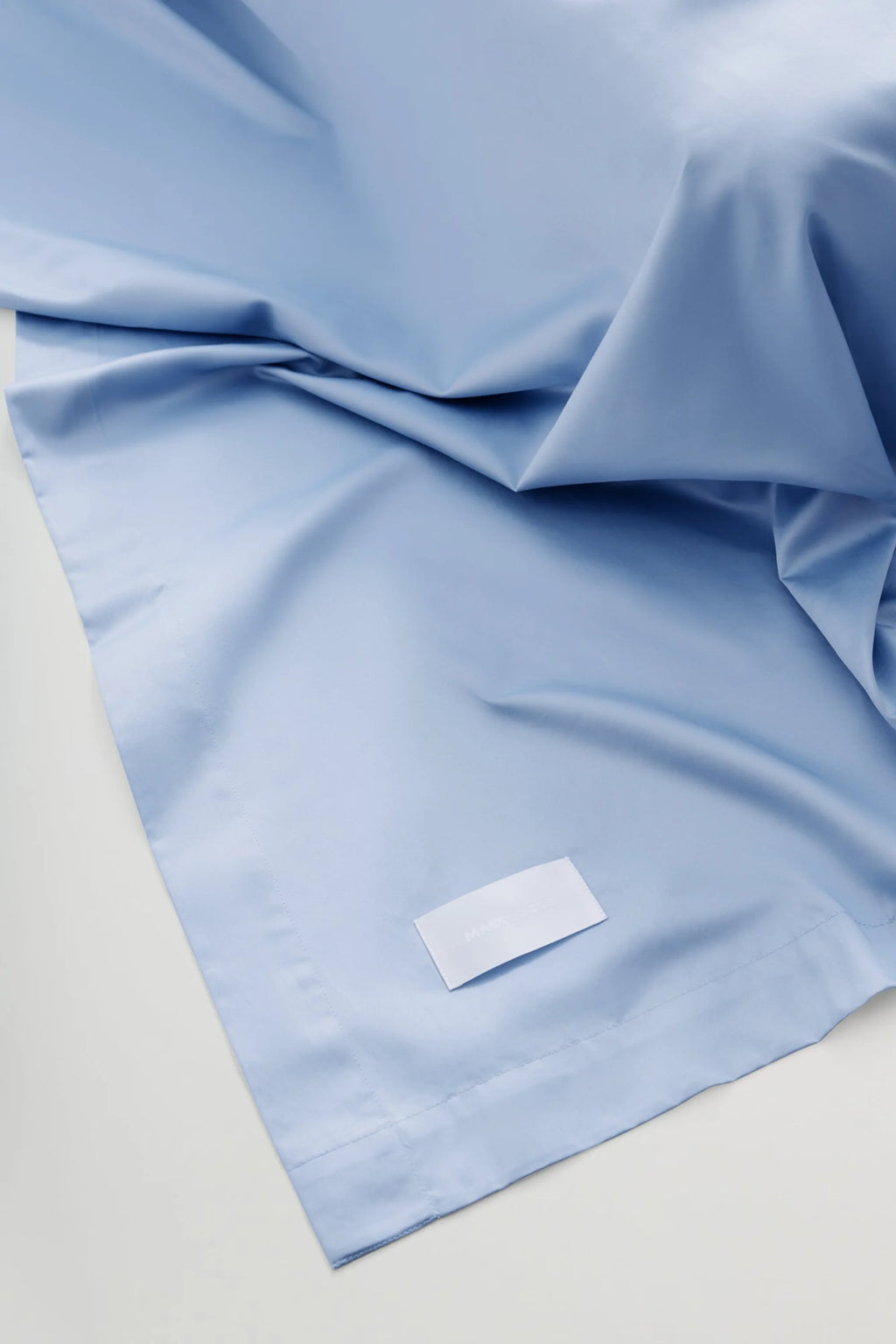 Magniberg Mother Sateen Flat Sheet, a luxurious light blue sateen flat sheet crafted from smooth, soft cotton sateen with a subtle sheen. The crisp yet fluid fabric drapes elegantly, showcasing gentle folds and a clean, minimalist design. Ideal for modern minimalist bedrooms, adding a touch of contemporary luxury and comfort.