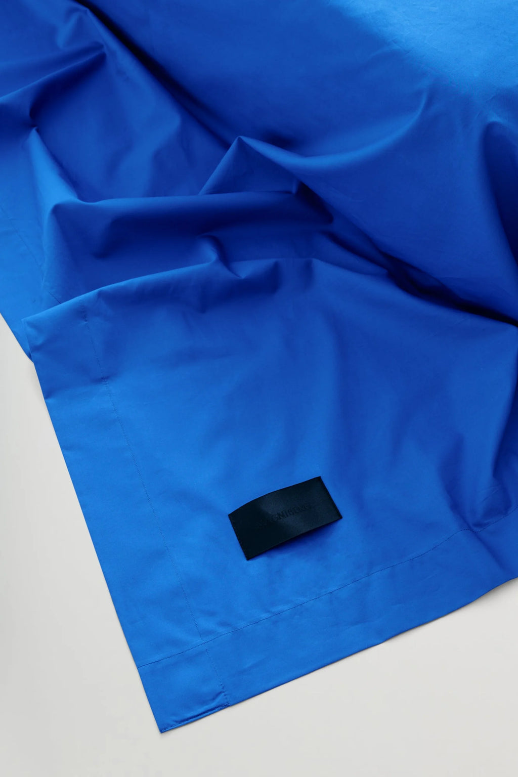 Magniberg Mother Poplin Flat Sheet, a vibrant royal blue flat sheet crafted from crisp cotton poplin fabric, featuring a smooth texture and elegant soft folds. This luxurious cobalt blue bedding piece includes a subtle black brand label sewn onto the corner. Perfect for adding a bold color statement to modern minimalist bedrooms or contemporary sleeping spaces.