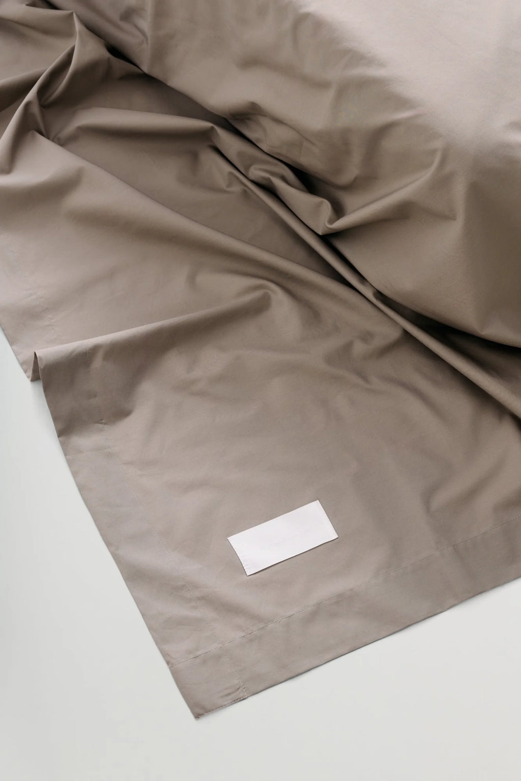 Magniberg Mother Poplin Flat Sheet, a luxurious taupe cotton poplin flat sheet featuring a smooth, crisp texture and soft, organic folds. This premium greige bedding is perfect for creating a modern minimalist or Scandinavian aesthetic in contemporary bedrooms.