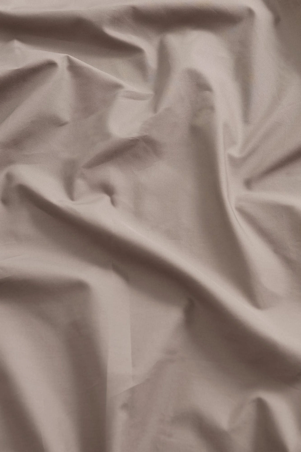 Magniberg Mother Poplin Flat Sheet, a luxurious and crisp beige cotton poplin flat sheet with soft wrinkles creating subtle texture and shadow. This neutral greige bed linen offers a smooth, matte finish, embodying a minimalist and contemporary Scandinavian design aesthetic, ideal for creating a serene and stylish bedroom or modern minimalist interiors.
