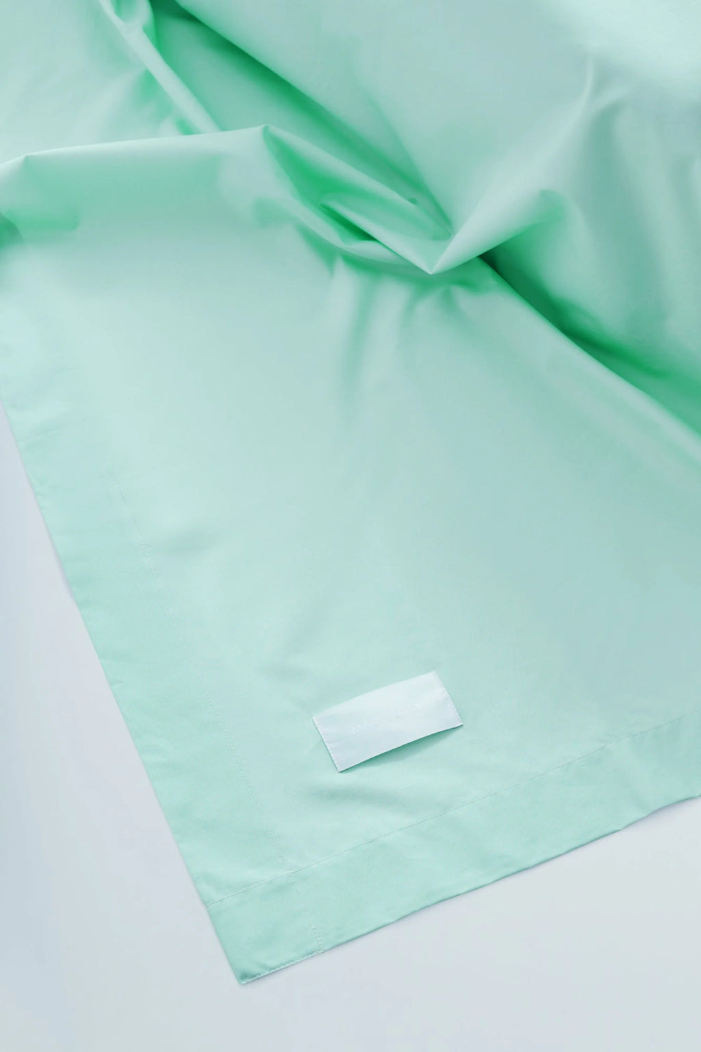 Magniberg Mother Poplin Flat Sheet, a crisp mint green poplin cotton flat sheet with a smooth, matte finish and subtle folds, featuring neatly hemmed edges. This pastel green bedding is perfect for creating a modern minimalist bedroom or a clean, contemporary sleeping space.