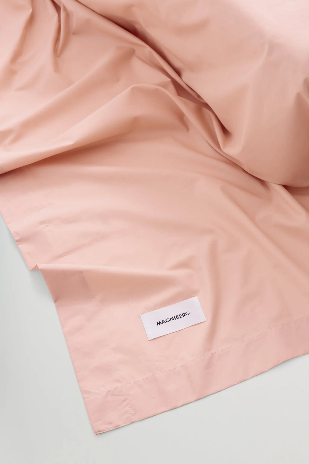Magniberg Mother Poplin Flat Sheet, a luxurious and crisp flat sheet crafted from premium 100% cotton poplin in a soft, muted blush pink color. This smooth, breathable bedding drapes elegantly, offering a refined texture and modern minimalist aesthetic. Ideal for creating a serene Scandinavian bedroom or adding a touch of contemporary elegance to your bed.