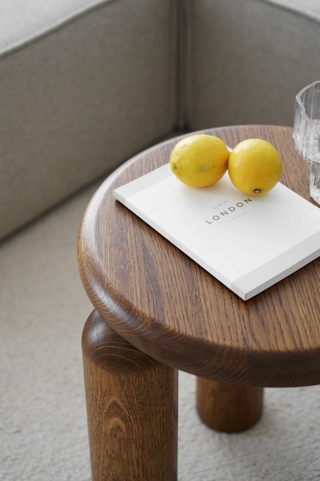 TAMO Neo Side Table, a close-up lifestyle shot of the premium solid dark oak wood with visible grain patterns on its round top and cylindrical legs, holding two lemons and a book. Ideal for modern living spaces.