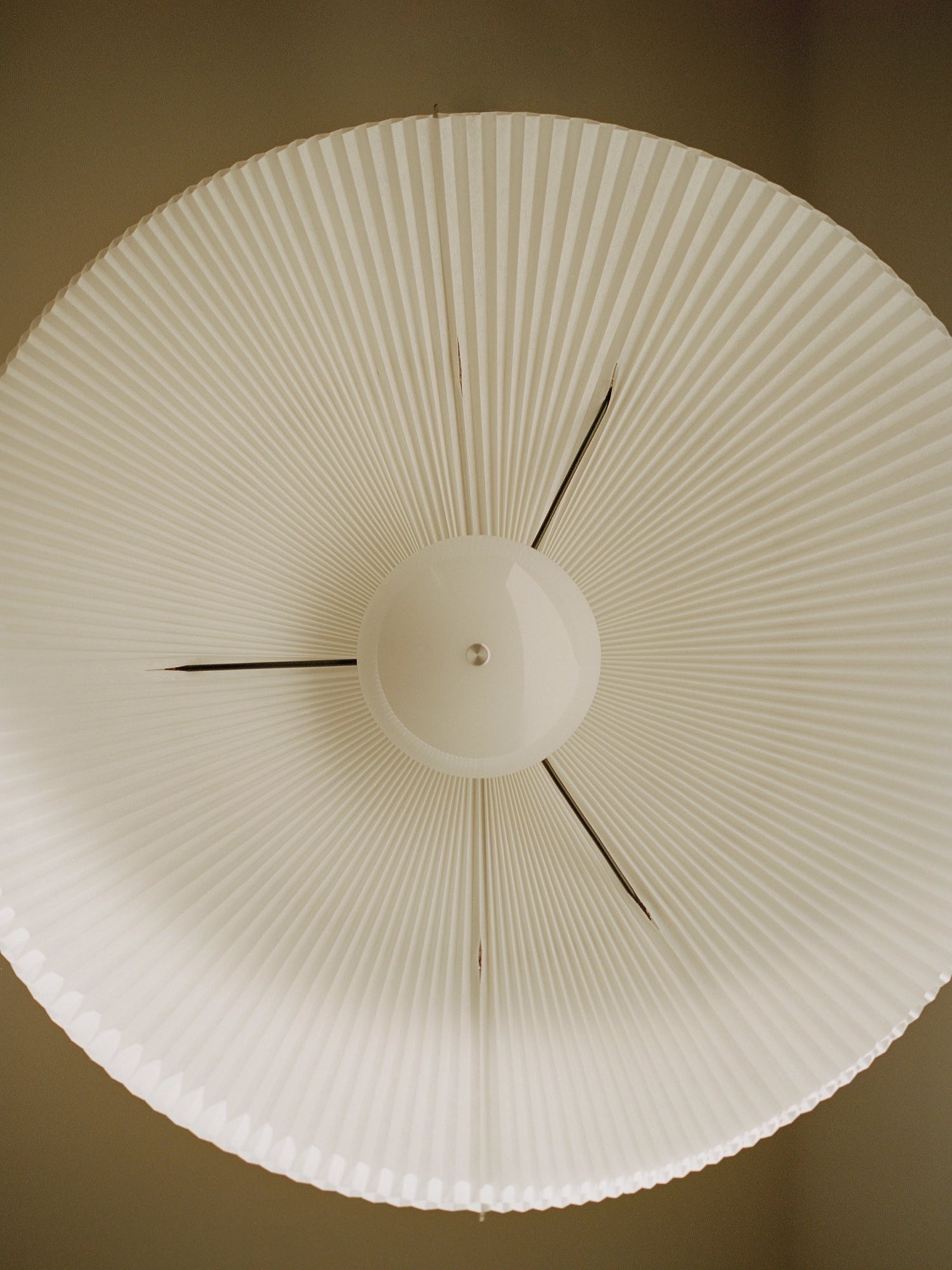 New Works Nebra Pendant Lamp, a modern minimalist pendant lamp featuring a wide, circular pleated paper shade in a soft cream color, emanating from a smooth central white dome. Thin dark metal rods provide structural support, creating a subtle, radial pattern. This Scandinavian-inspired light fixture is perfect for adding soft, diffused lighting to contemporary living rooms, dining areas, or bedrooms.
