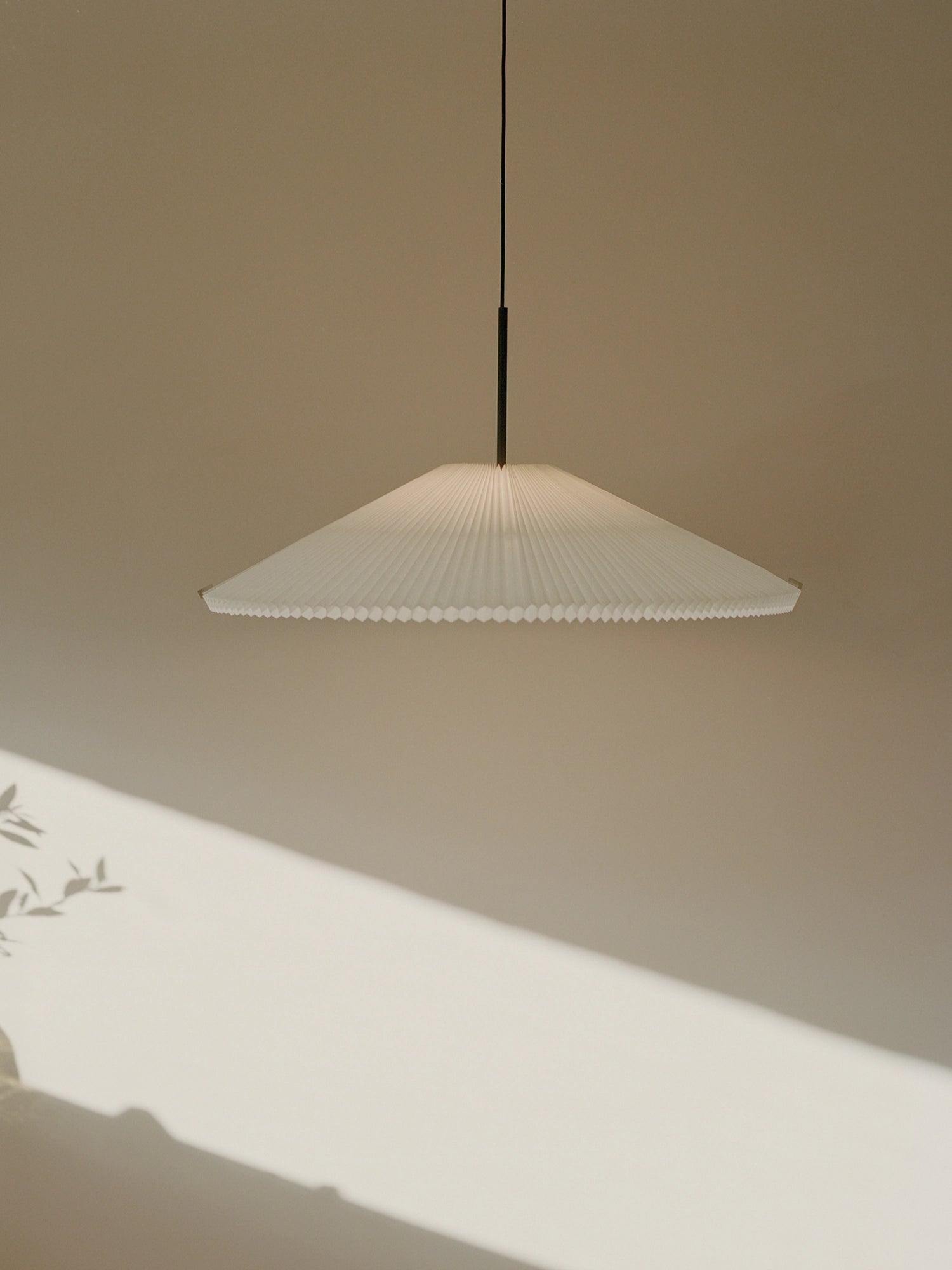 New Works Nebra Pendant Lamp, a minimalist Scandinavian ceiling light featuring a wide, shallow conical shade crafted from delicately pleated white paper with a distinct corrugated edge, suspended by a sleek black cord and metal accent. This elegant pleated paper lamp provides soft, diffused light, perfect for modern living rooms, japandi dining areas, or serene bedroom interiors.