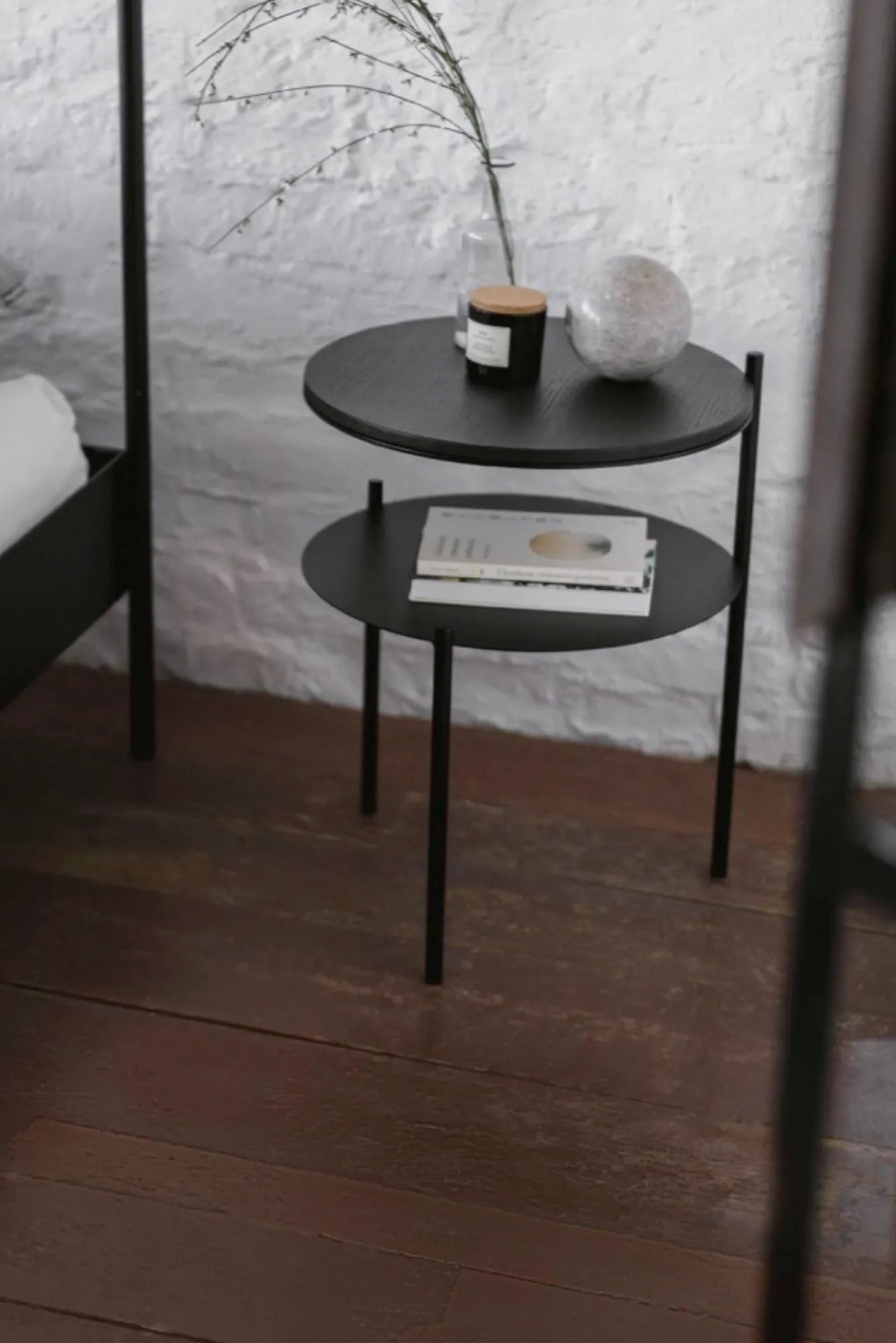 noo.ma Tu Bedside Table, a sleek modern minimalist side table featuring two round shelves crafted from black wood with a visible grain, supported by a slender matte black metal frame and three minimalist cylindrical legs. This contemporary two-tier nightstand offers ample surface area for bedside essentials and is ideal for Scandinavian bedrooms, small apartment living rooms, or any minimalist interior seeking a functional and stylish black end table.