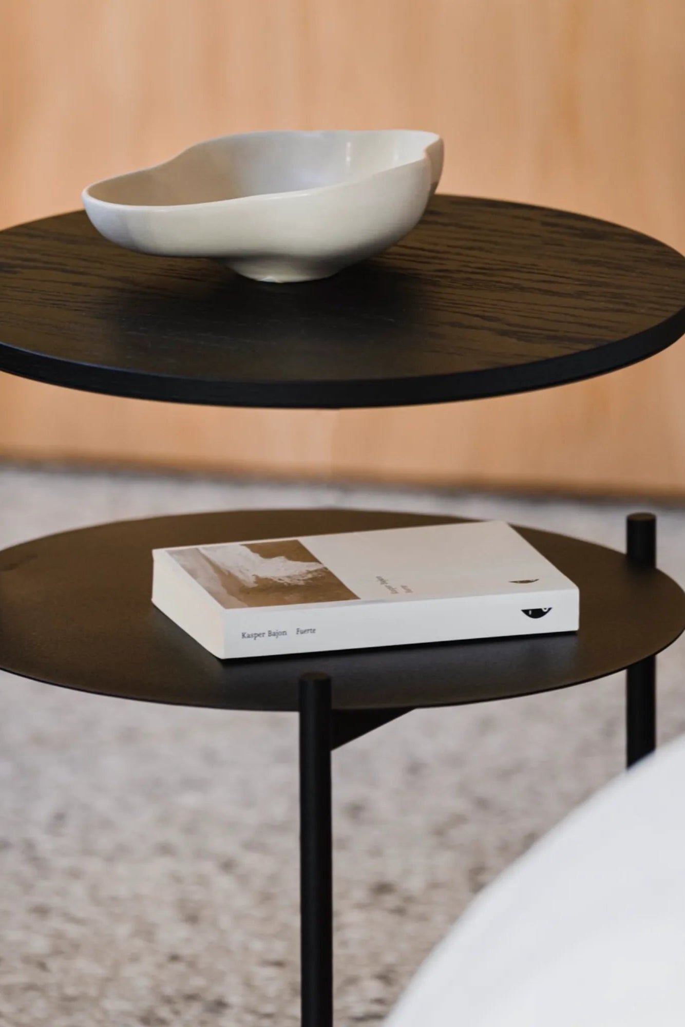 noo.ma Tu Bedside Table, a sleek modern minimalist two-tier side table featuring a dark black oak wood top shelf with visible grain and a complementary matte black metal lower shelf and slender cylindrical legs. Its clean lines and round form create a contemporary Scandinavian aesthetic. Ideal as a stylish bedside table for bedrooms or a versatile accent table in living rooms and entryways.