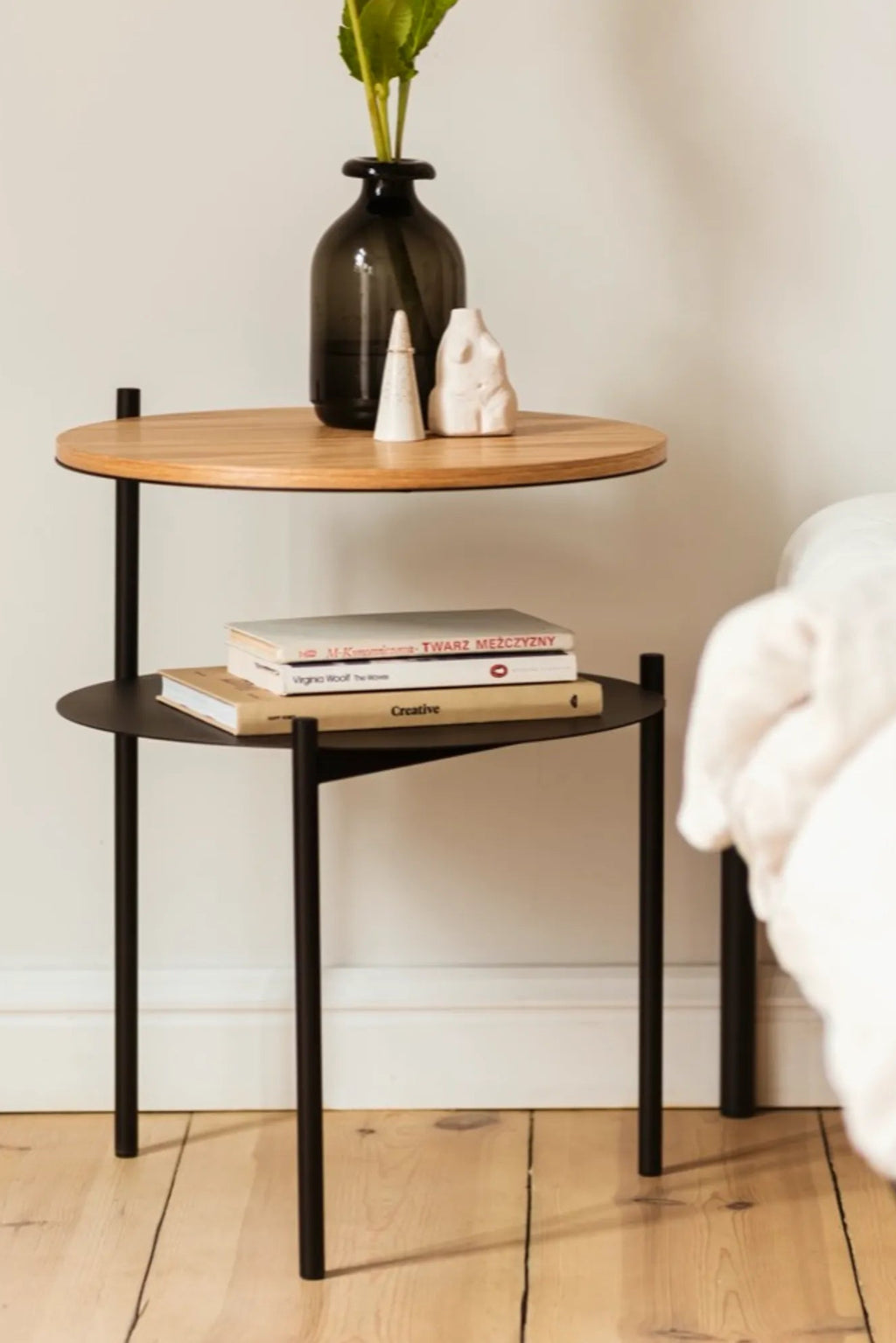 noo.ma Tu Bedside Table, a modern minimalist two-tiered side table featuring a natural light wood round top shelf and a contrasting matte black metal lower shelf. Its contemporary design includes slender black metal legs that elegantly support the offset circular shelves, creating a sleek and functional silhouette. Ideal as a Scandinavian bedside table, a minimalist nightstand, or a small accent table for modern living rooms and compact spaces.