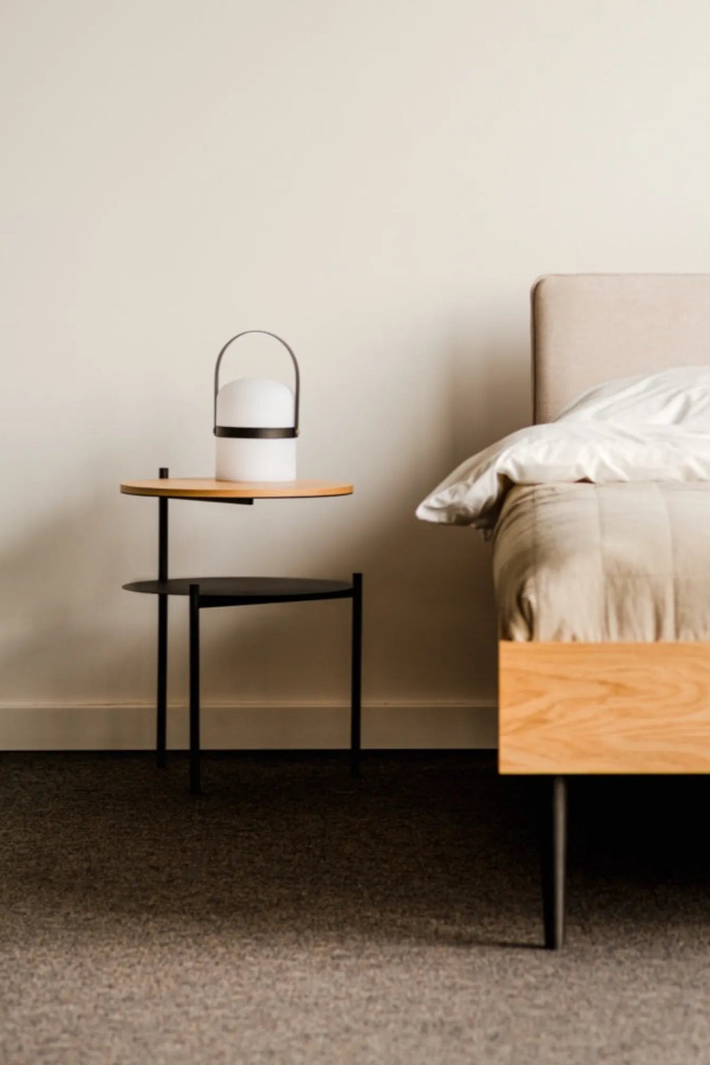 noo.ma Tu Bedside Table, a modern minimalist two-tier side table featuring a round natural light wood top shelf and a lower matte black metal shelf, supported by a sleek black metal frame. This Scandinavian-inspired nightstand offers clean lines and functional design for contemporary bedrooms or small living room accents.