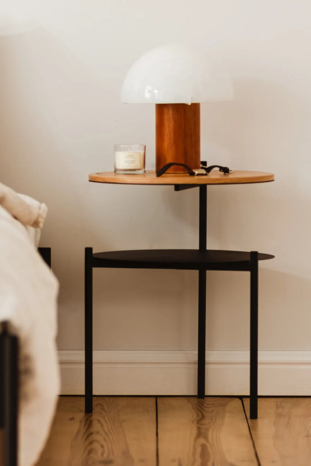 noo.ma Tu Bedside Table, a modern minimalist two-tier side table featuring a sleek matte black metal frame with an upper round natural wood shelf and a lower round black metal shelf. This contemporary design offers functional storage with clean lines, blending natural wood tones with industrial chic elements. Perfect as a Scandinavian bedside table for bedrooms or a stylish round end table for small living room spaces.