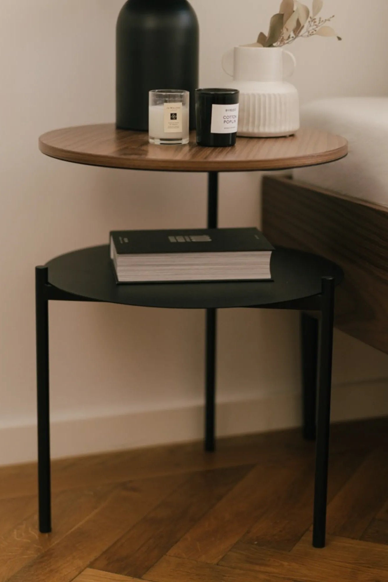 noo.ma Tu Bedside Table, a modern minimalist two-tier side table featuring a round dark walnut wood veneer top shelf contrasted with a matte black metal lower shelf and sleek cylindrical frame.