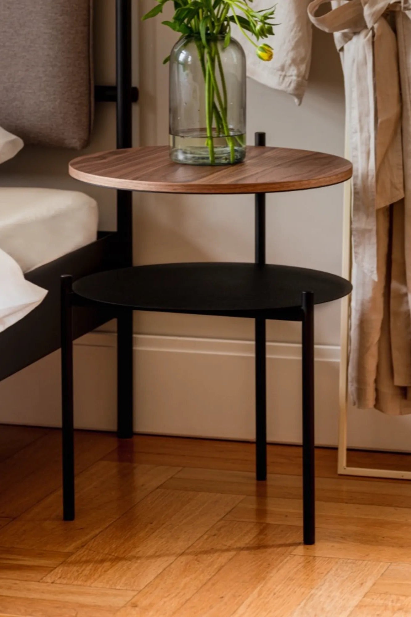 noo.ma Tu Bedside Table, a modern minimalist two-tier side table featuring a round dark walnut wood veneer top and a lower round matte black metal shelf, supported by slim black metal legs. This Scandinavian-inspired nightstand is ideal for contemporary bedroom decor or as a functional accent table in small living spaces.