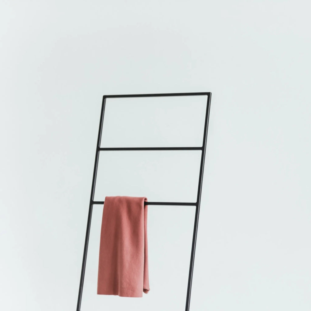 noo.ma Tows Ladder, a sleek matte black metal towel ladder featuring a modern minimalist lean-to design with multiple rectangular rungs, elegantly draped with a dusty rose-colored towel. This versatile freestanding rack is perfect for Scandinavian bathrooms, industrial chic bedrooms, or contemporary living rooms for blanket storage.