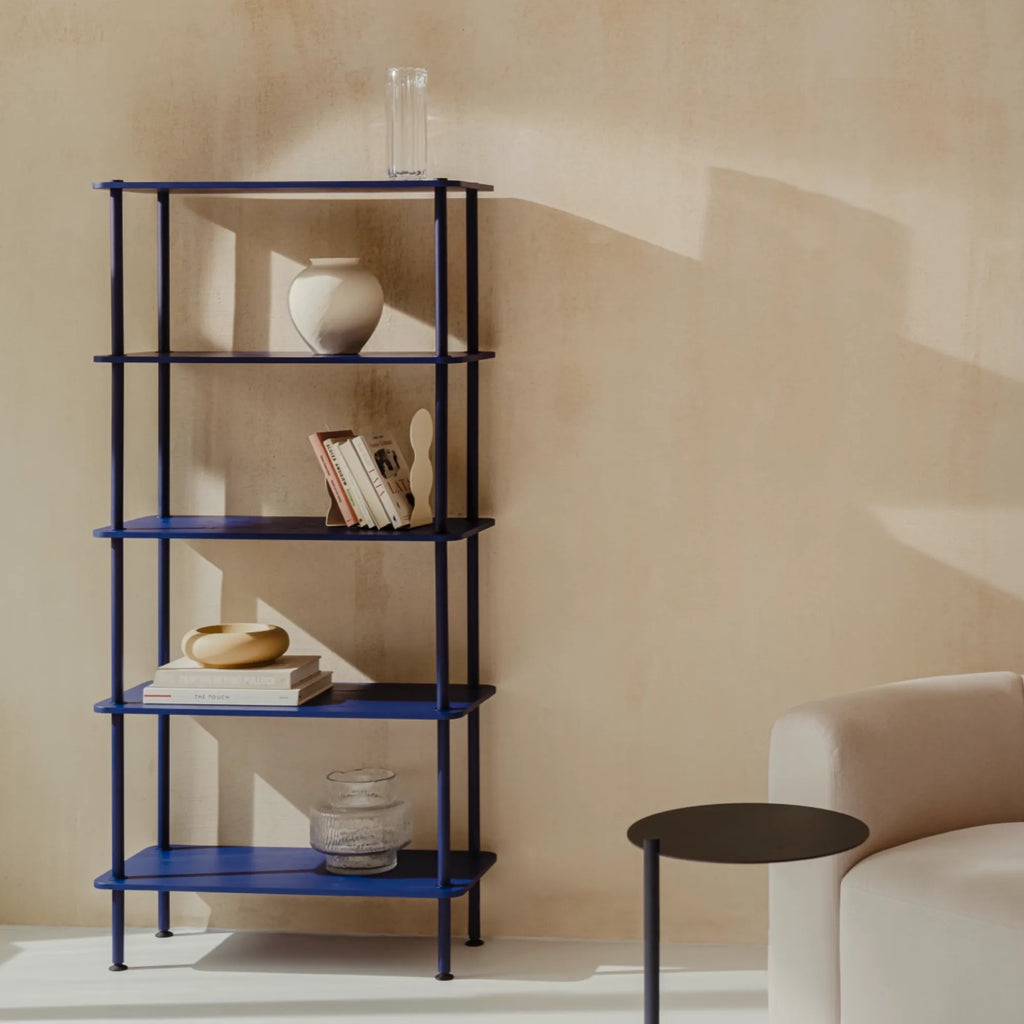 noo.ma Teidi Shelving Unit 5-tier, a striking deep blue matte metal shelving unit with a minimalist, contemporary design featuring five open rectangular tiers and slender cylindrical posts. This versatile 5-tier blue metal bookcase offers ample display and storage, perfect for adding a bold pop of color to modern living rooms, home offices, or Scandinavian-inspired interiors.