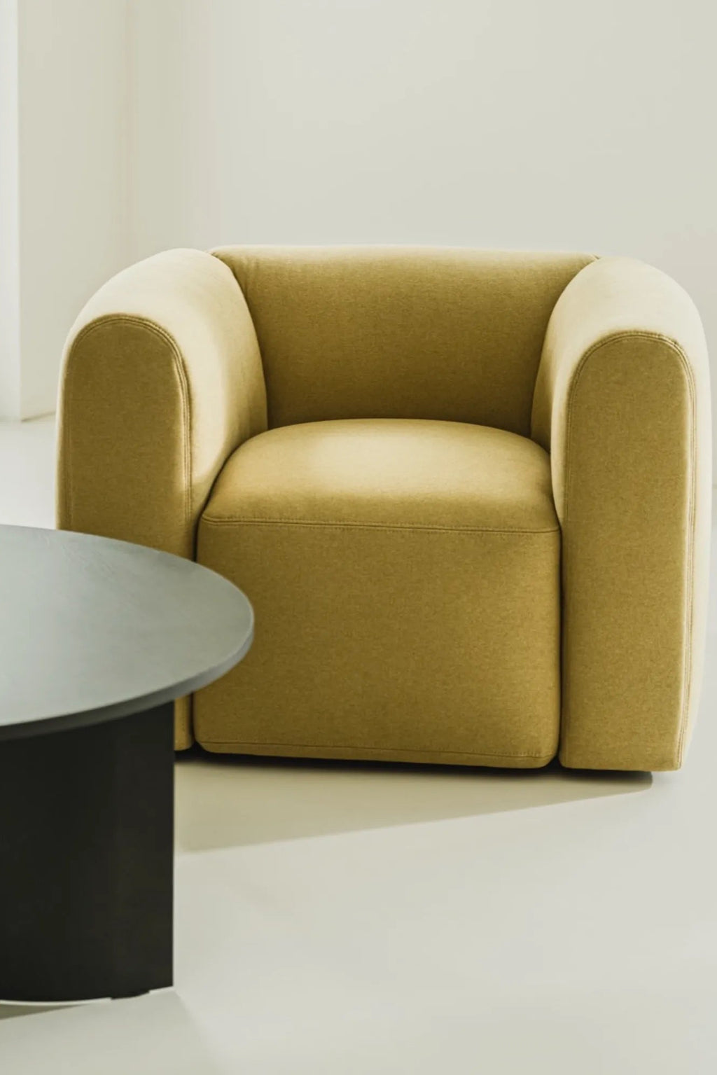 noo.ma Flom Armchair, a contemporary organic modern lounge chair upholstered in a rich mustard yellow textured fabric with a plush, rounded silhouette and curvaceous design. This minimalist accent chair features thick, rolled arms and a deep seat, offering an inviting and sculptural form. Perfect as a statement piece for a modern living room, cozy reading nook, or chic bedroom accent.