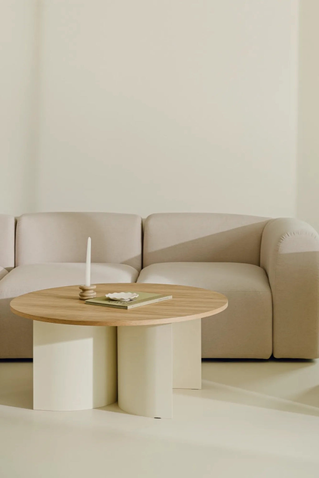 noo.ma Gavo Coffee Table, a modern minimalist round coffee table featuring a light natural wood tabletop, such as oak or ash, with a unique sculptural base composed of curved cream-colored matte elements. This organic modern design is ideal for contemporary living rooms seeking a touch of Scandinavian elegance.