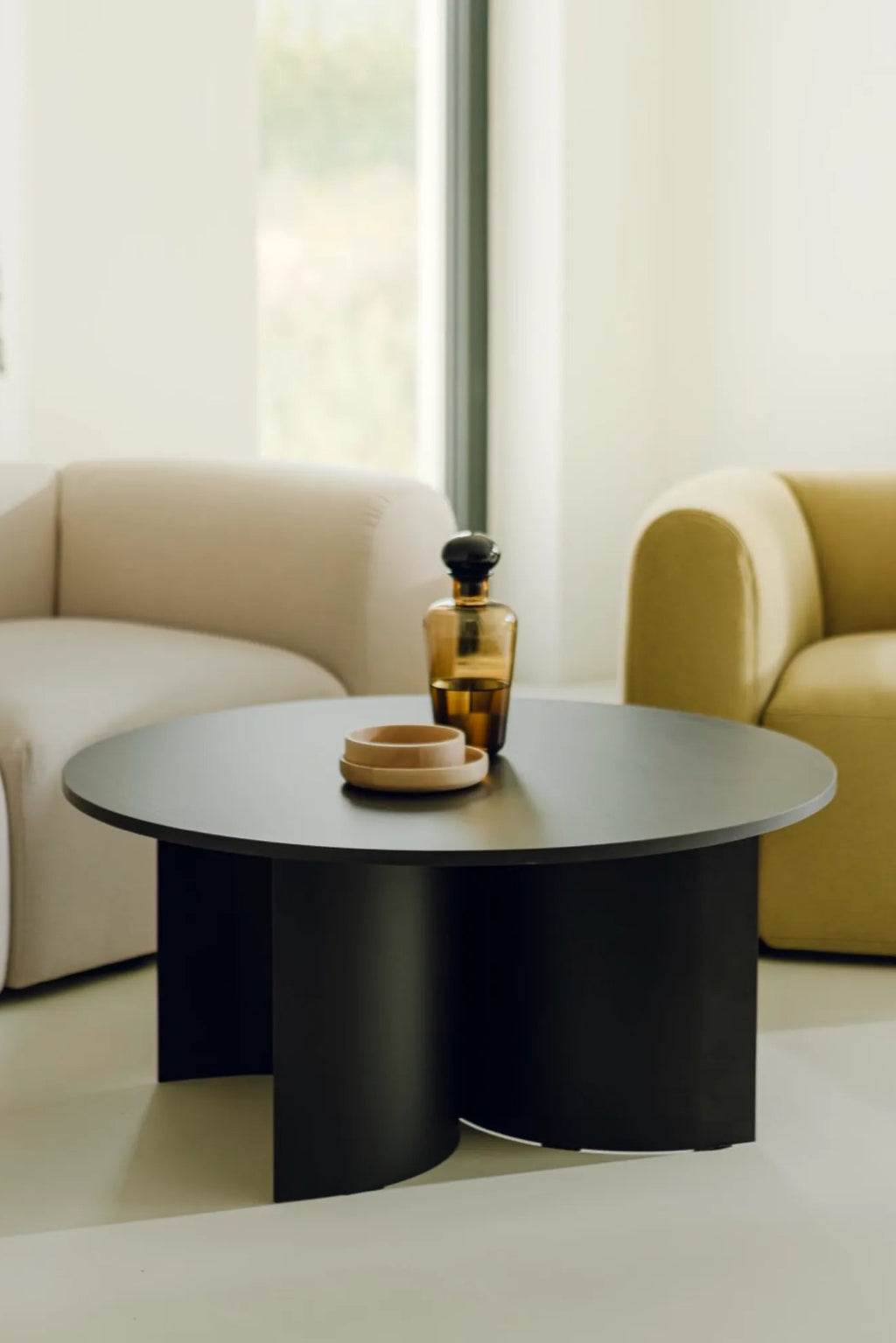 noo.ma Gavo Coffee Table, a modern minimalist round coffee table featuring a smooth matte black finish and a distinctive sculptural base with three curved, cylindrical legs. This contemporary black coffee table offers an elegant focal point for stylish living rooms and organic modern interiors.
