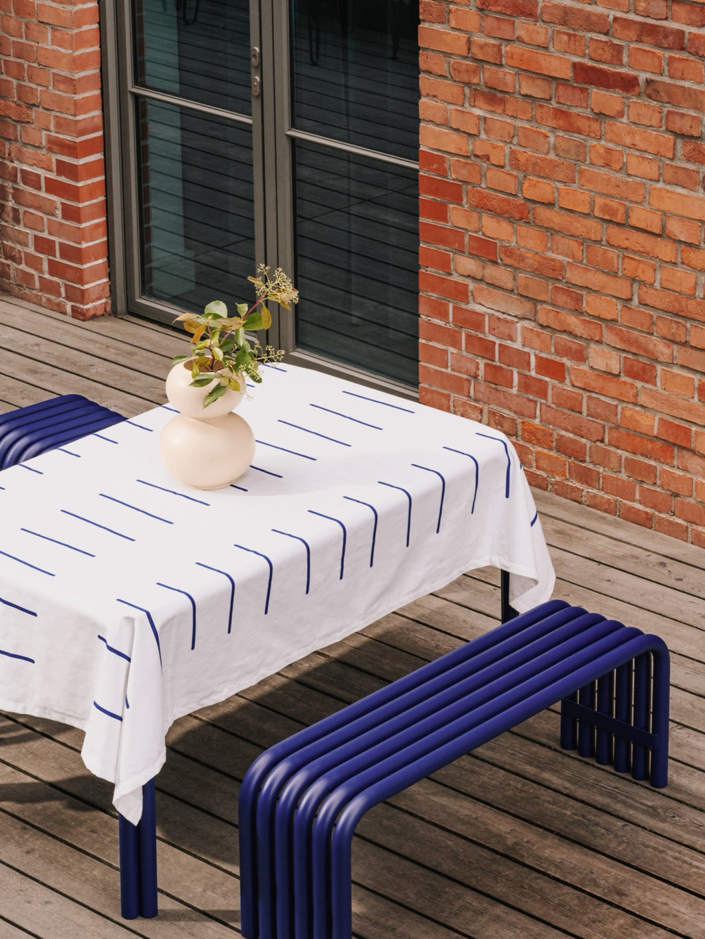 noo.ma Nokk Bench, a vibrant deep blue tubular metal bench featuring a minimalist, curved design and a smooth, powder-coated finish, creating a striking sculptural aesthetic. This contemporary outdoor bench is perfect for modern patios, garden seating, or as a bold dining bench in design-conscious spaces.