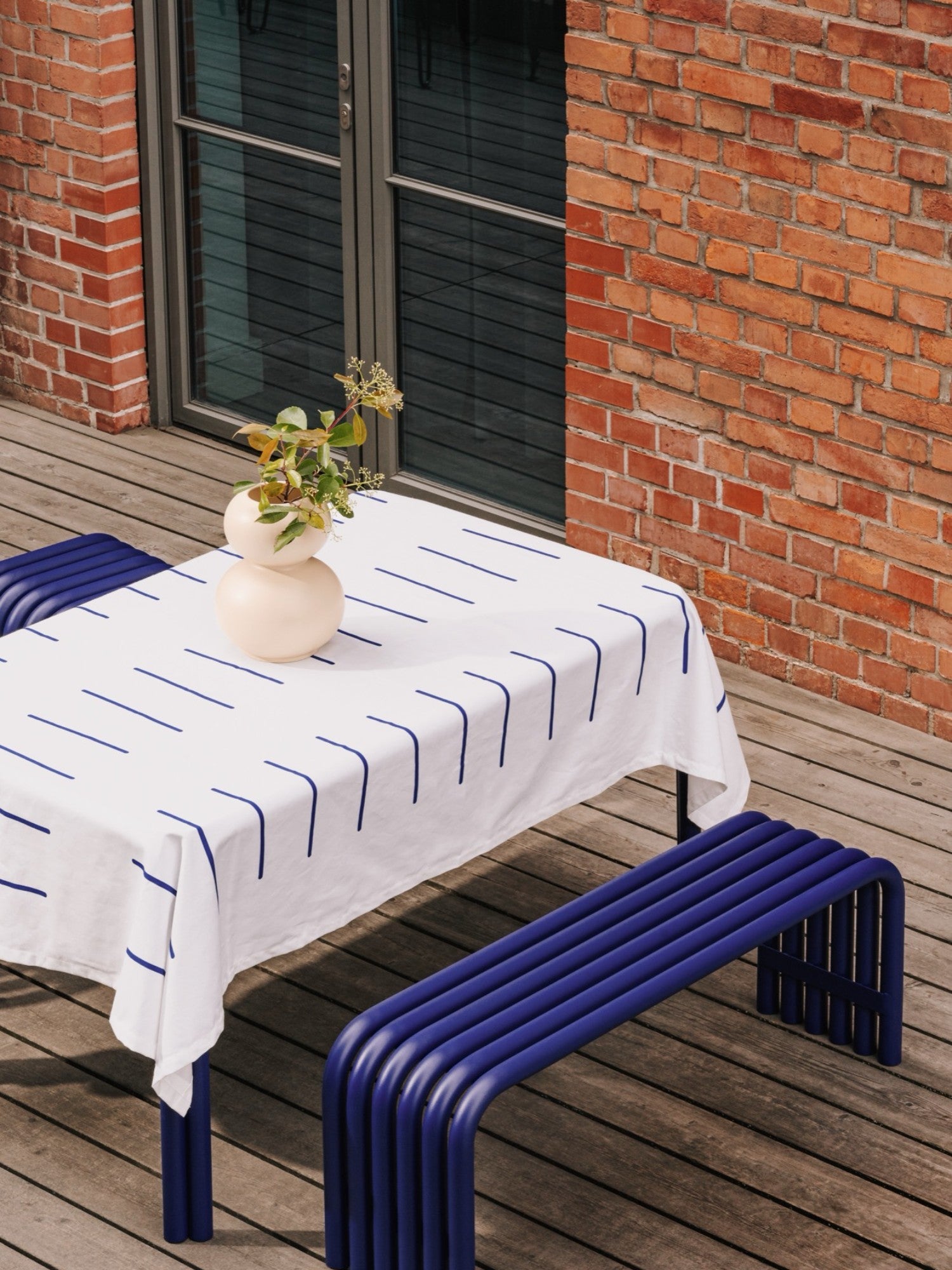 noo.ma Nokk Bench, a vibrant deep blue tubular metal bench featuring a minimalist, curved design and a smooth, powder-coated finish, creating a striking sculptural aesthetic. This contemporary outdoor bench is perfect for modern patios, garden seating, or as a bold dining bench in design-conscious spaces.