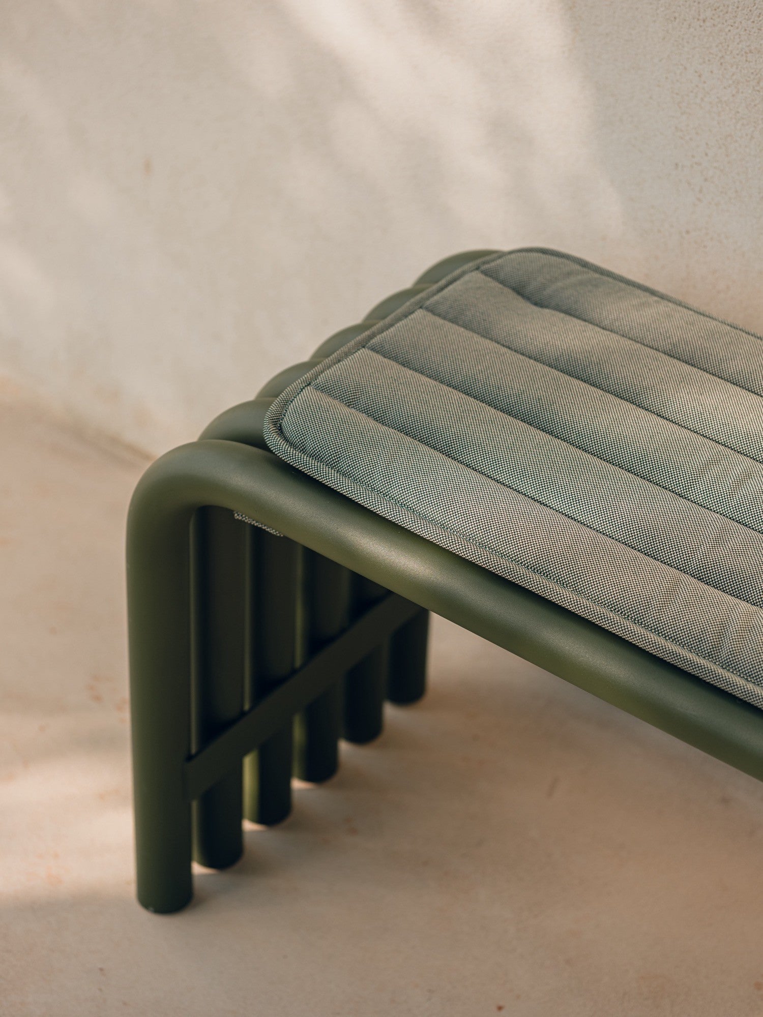 noo.ma Nokk Bench, a modern minimalist outdoor bench featuring a dark olive green matte tubular metal frame with rounded legs and a light sage green removable cushion.