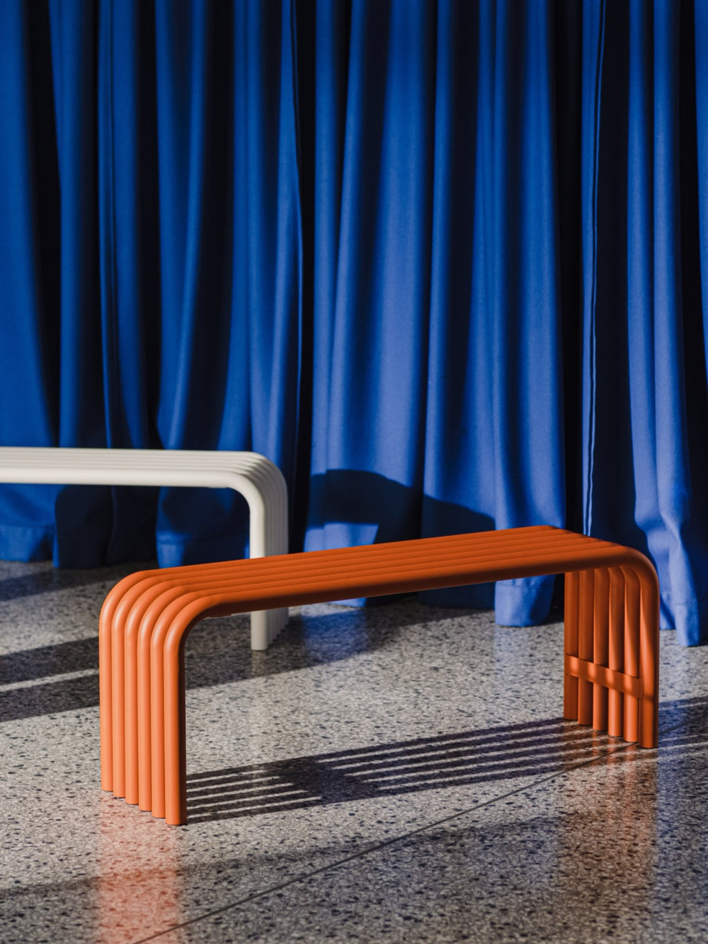 noo.ma Nokk Bench, a terracotta orange powder-coated metal bench featuring a distinctive tubular design with parallel rounded steel bars forming the seat and legs, creating a ribbed, sculptural profile.