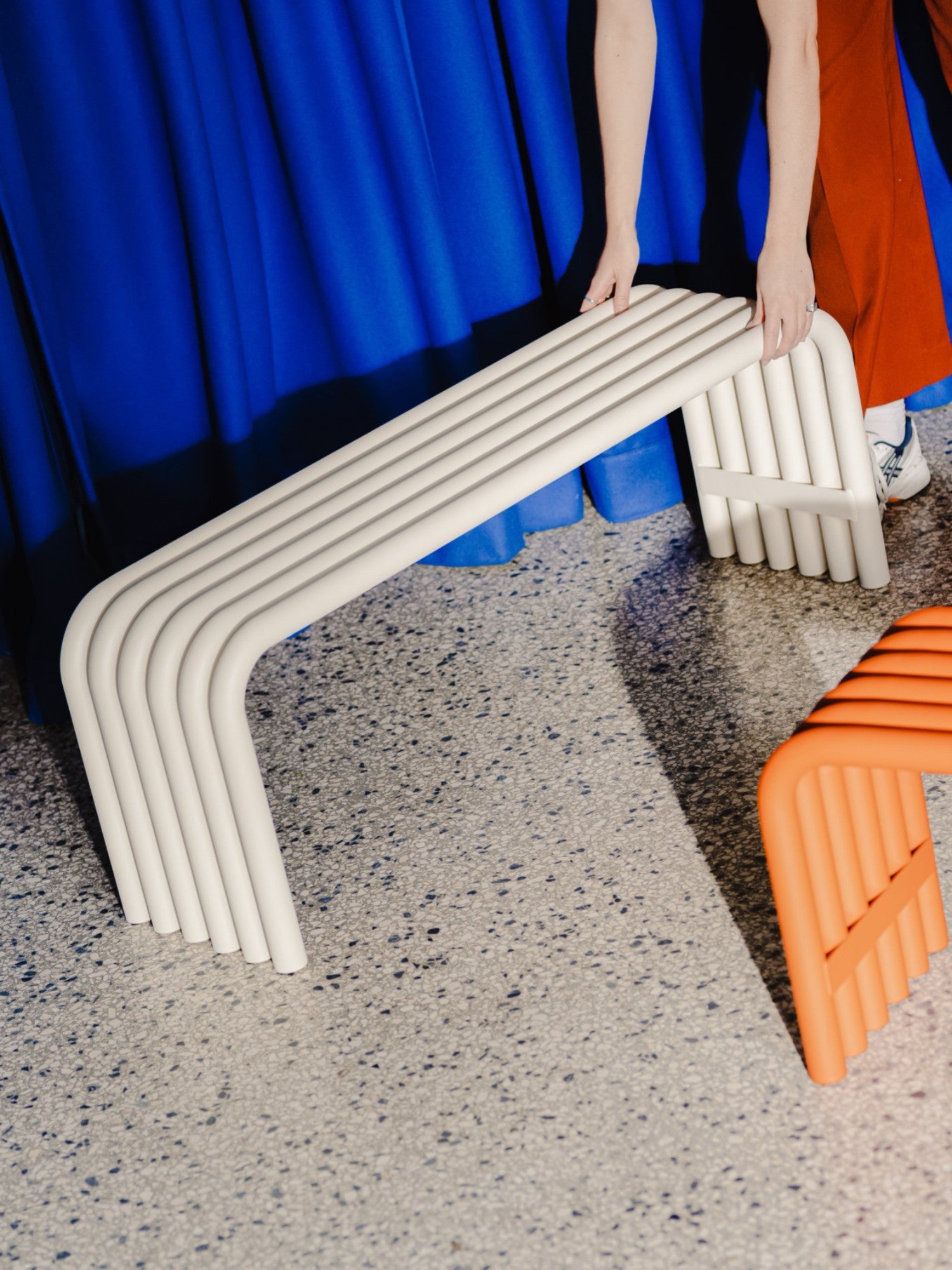 noo.ma Nokk Bench, a contemporary sculptural bench crafted from light beige powder-coated metal, featuring a distinctive tubular design with parallel curved elements forming the seat and inverted U-shaped legs.