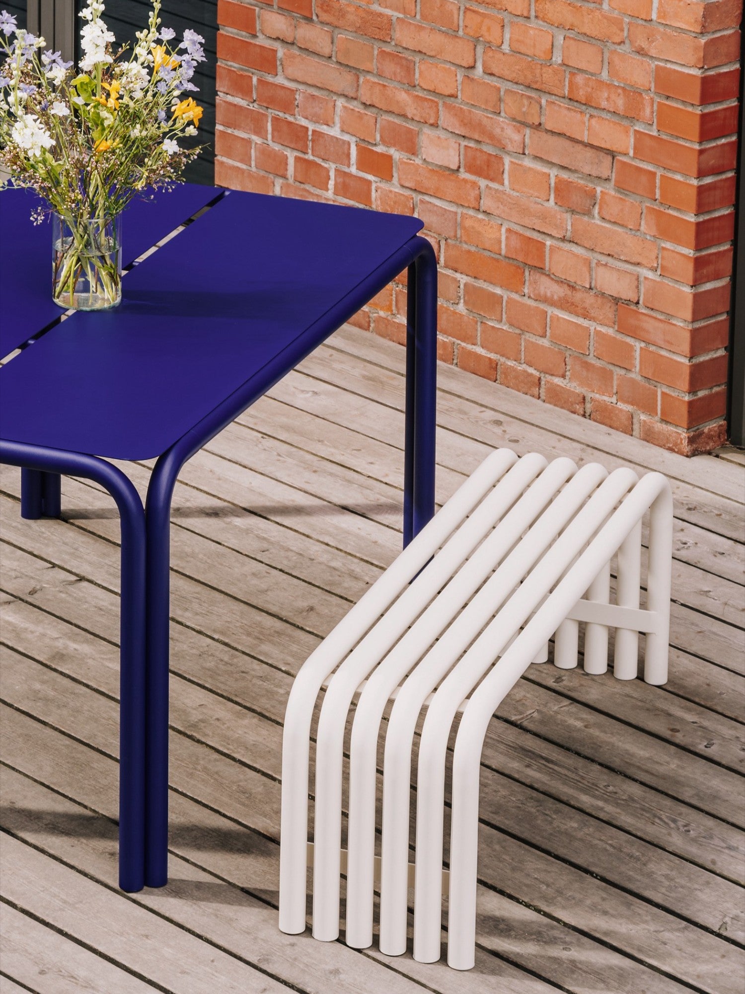 noo.ma Nokk Bench, a modern minimalist outdoor bench crafted from durable white powder-coated tubular metal with a series of gracefully curved slats forming a comfortable and stylish seat. Its clean, contemporary design and weather-resistant finish make it perfect for patio seating, garden accents, or balcony arrangements.