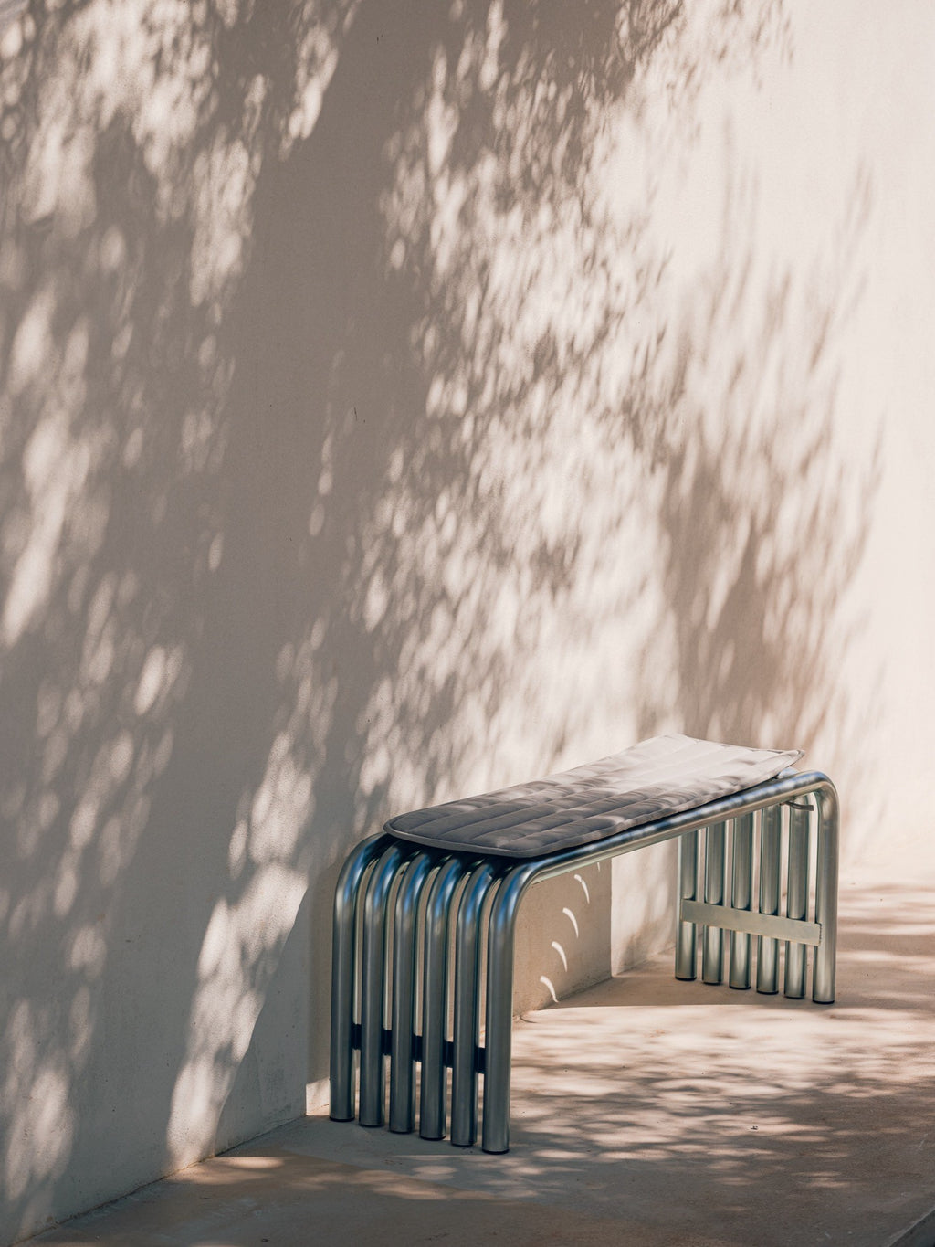 noo.ma Nokk Bench, a sleek modern minimalist bench featuring a distinctive frame crafted from polished chrome metal tubing, forming a sculptural, industrial-chic base. It is topped with a dark grey upholstered cushion, offering a comfortable contrast to the metallic structure. This contemporary metal bench is ideal for modern entryways, outdoor patios, or minimalist living spaces.