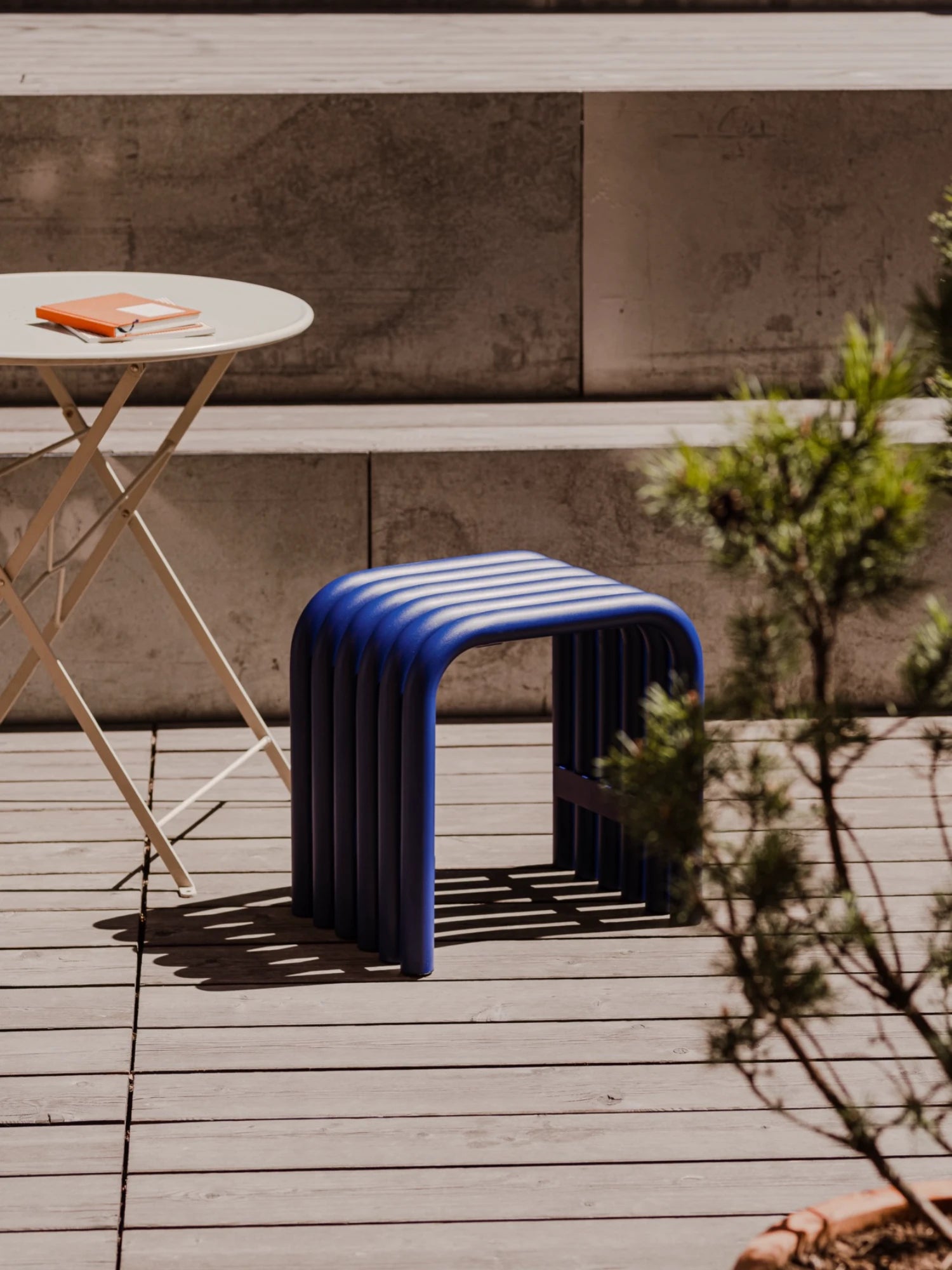 A dark blue Nokk Stool with a bent-tube design is placed on a wooden deck. The stool casts a striped shadow in the sunlight. A small round table with a notebook on it is next to the stool, with stone steps and a potted plant in the background.
