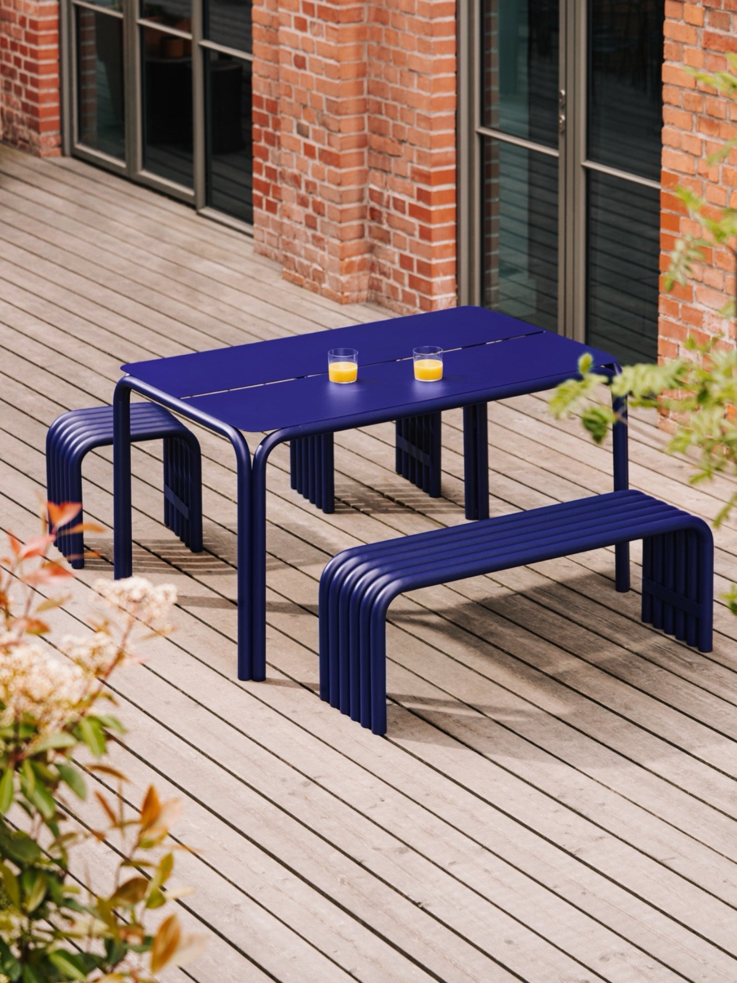 noo.ma Nokk Stool, a modern minimalist outdoor bench crafted from vibrant deep blue powder-coated metal, featuring a sleek rectangular slatted top and distinctive curved, parallel tubular legs.