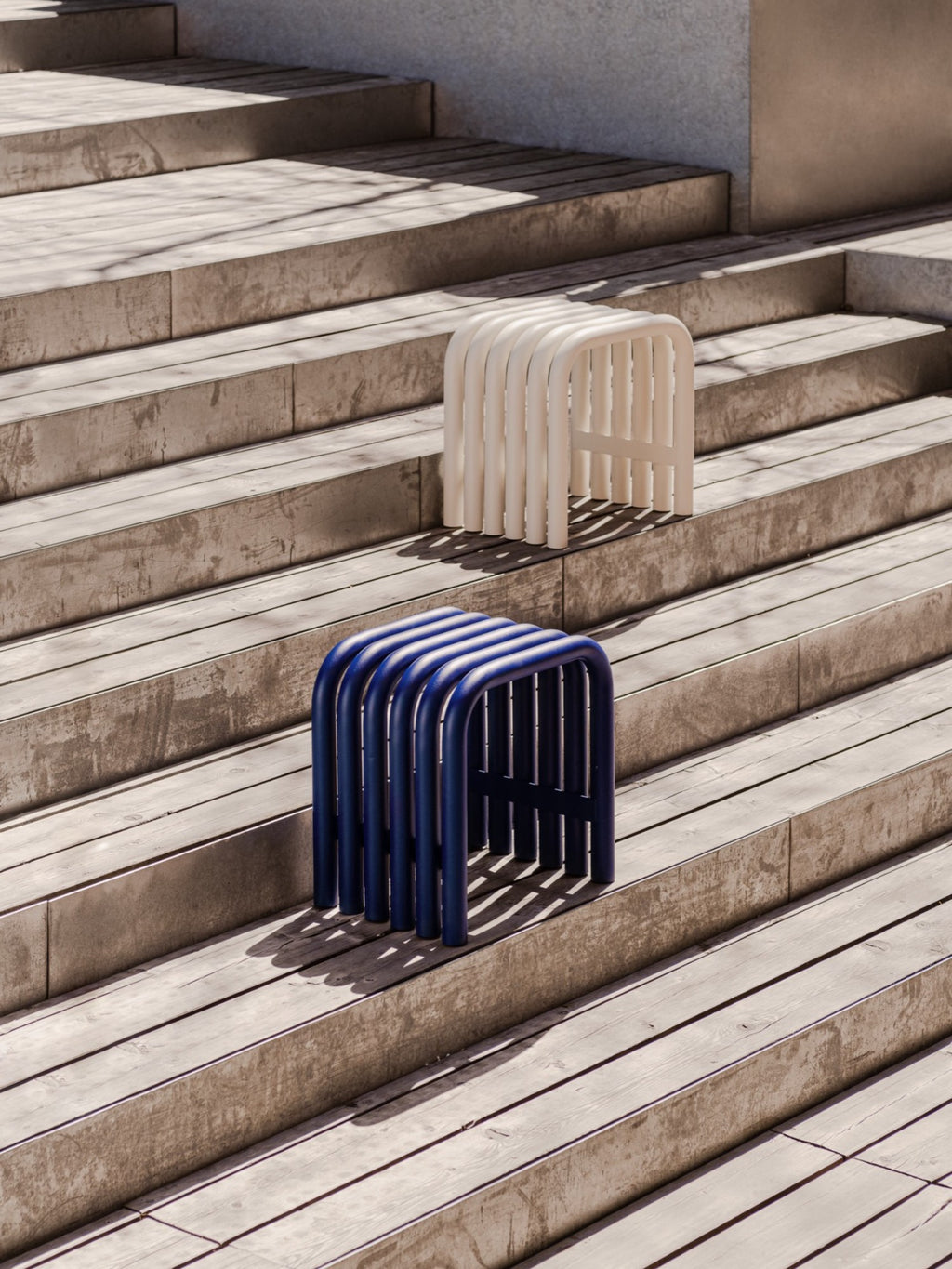 noo.ma Nokk Stool, a modern minimalist tubular metal stool presented in a striking dark blue and a soft off-white, featuring a distinctive U-shaped sculptural design. Crafted from durable powder-coated metal, its rounded edges and slatted construction offer a contemporary and playful aesthetic. This versatile accent stool or side table is ideal for modern living rooms, bedrooms, entryways, or as stylish indoor-outdoor furniture.