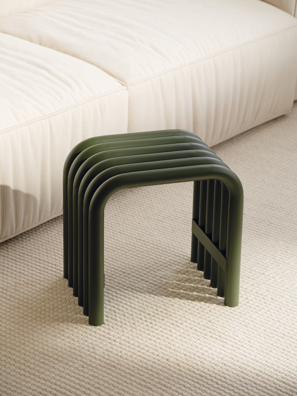 noo.ma Nokk Stool, a modern minimalist dark olive green tubular metal stool with a distinctive ribbed design and smooth matte finish. Crafted from powder-coated steel, its unique U-shaped profile creates a contemporary accent piece. Ideal as a stylish side table, extra seating, or a decorative plant stand for modern living rooms, bedrooms, or entryways.