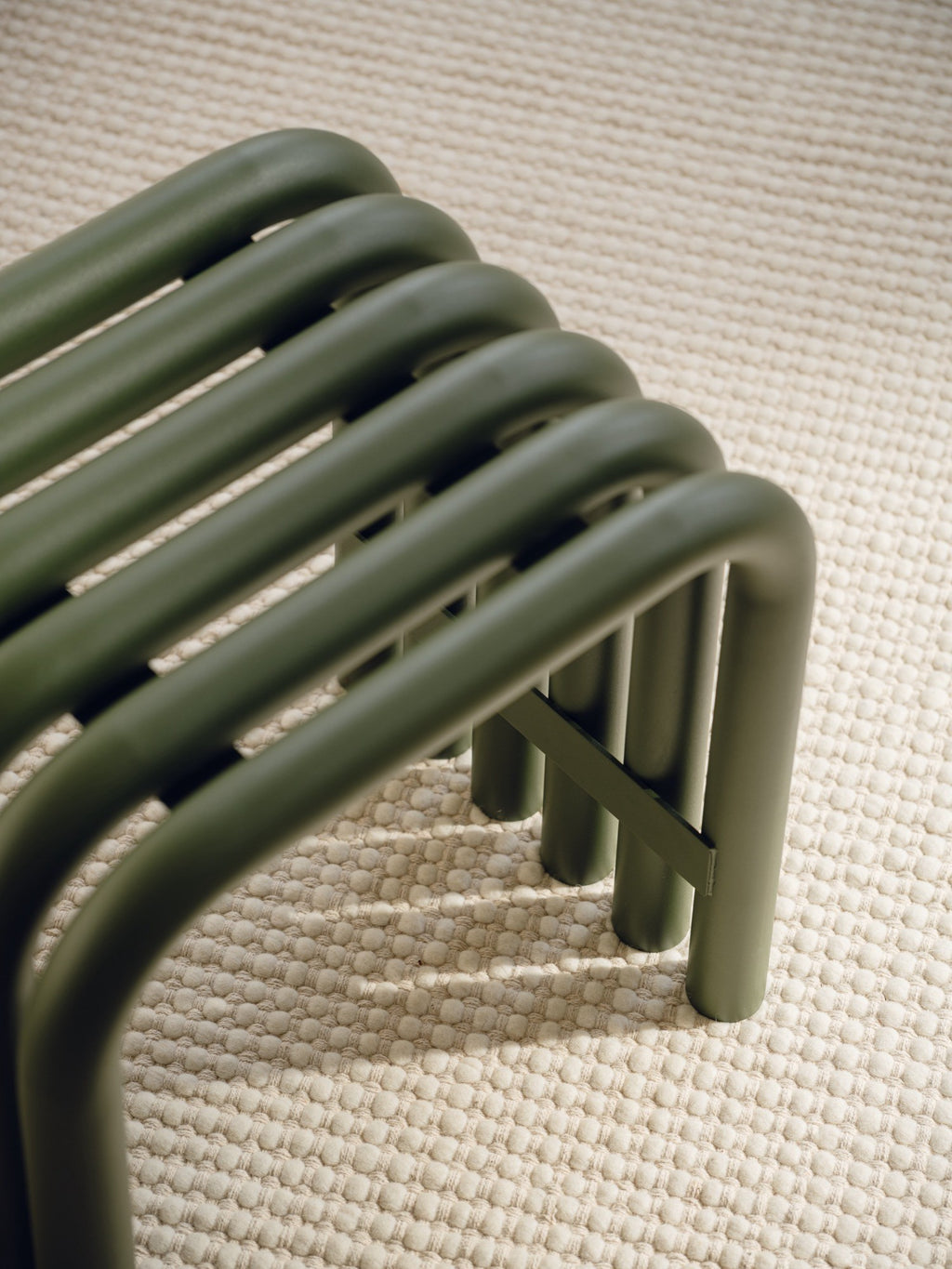 noo.ma Nokk Stool, a modern minimalist stool crafted from matte olive green tubular metal with a distinctive slatted seat and curved legs, showcasing a clean, contemporary design. This industrial chic metal bench provides versatile seating for an entryway, living room, or as a stylish bedroom accent, resting on a textured cream bobble rug.