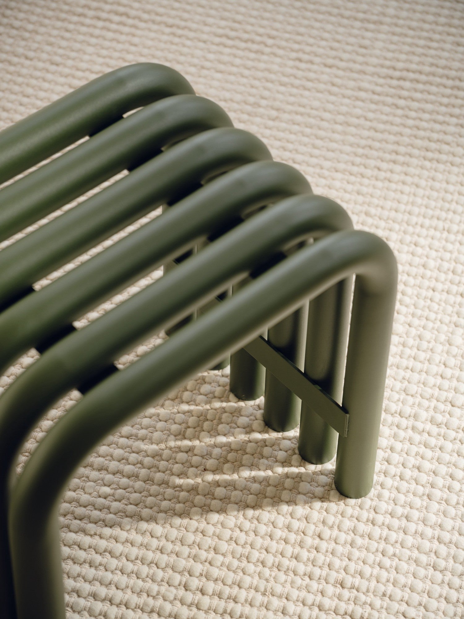 noo.ma Nokk Stool, a modern minimalist stool crafted from matte olive green tubular metal with a distinctive slatted seat and curved legs, showcasing a clean, contemporary design. This industrial chic metal bench provides versatile seating for an entryway, living room, or as a stylish bedroom accent, resting on a textured cream bobble rug.