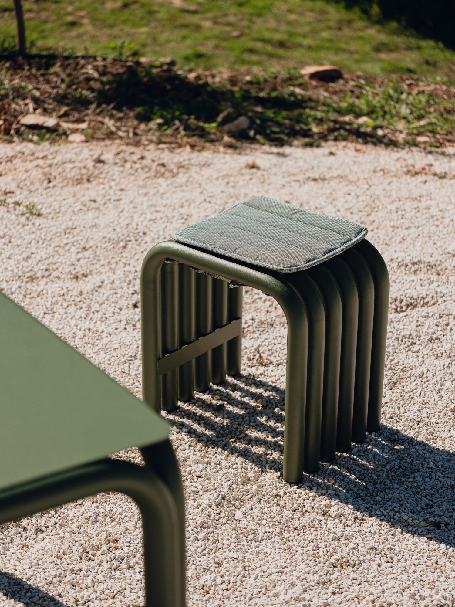 noo.ma Nokk Stool, a modern olive green metal stool featuring a unique base of curved, matte tubular steel legs and a soft grey-green padded fabric cushion. This contemporary accent stool is perfect for minimalist outdoor living spaces, patios, or as stylish garden furniture.