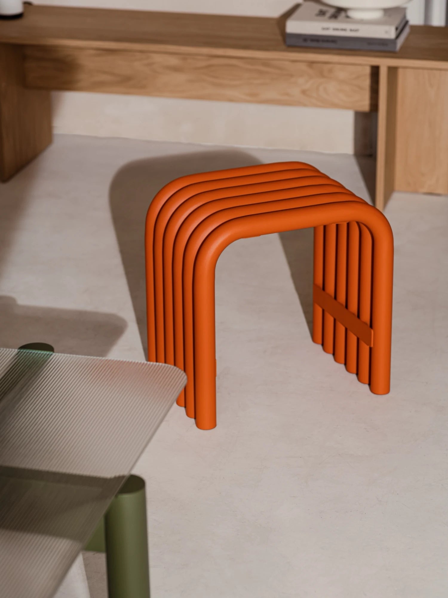 A minimalist Nokk Stool in a bright Orange Peel finish sits on a light-colored floor. The stool is made from a series of bent tubes and casts a distinct striped shadow. A portion of a dark blue upholstered chair is visible in the top right corner.