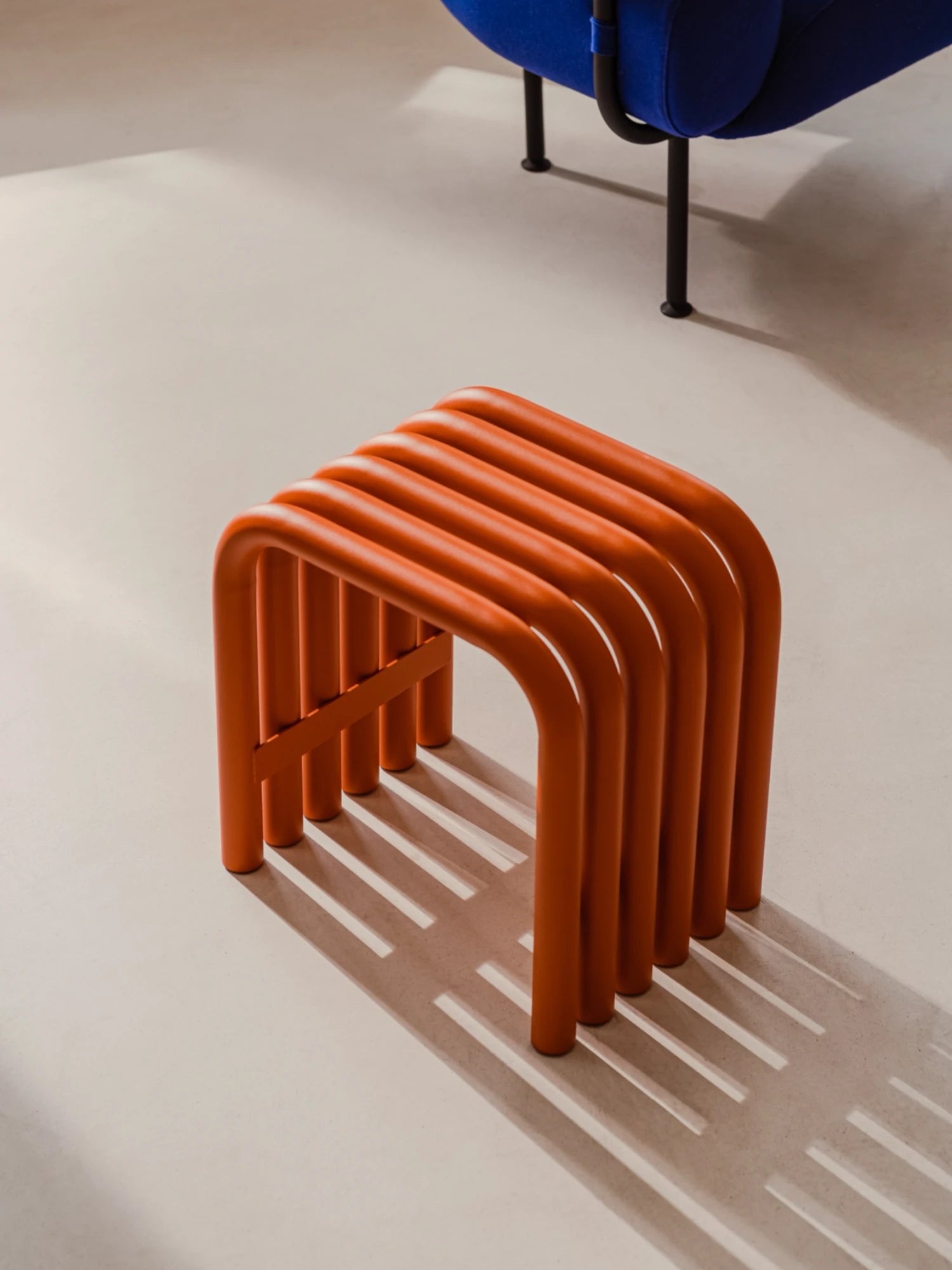 A minimalist Nokk Stool in a bright Orange Peel finish sits on a light-colored floor. The stool is made from a series of bent tubes and casts a distinct striped shadow. A portion of a dark blue upholstered chair is visible in the top right corner.
