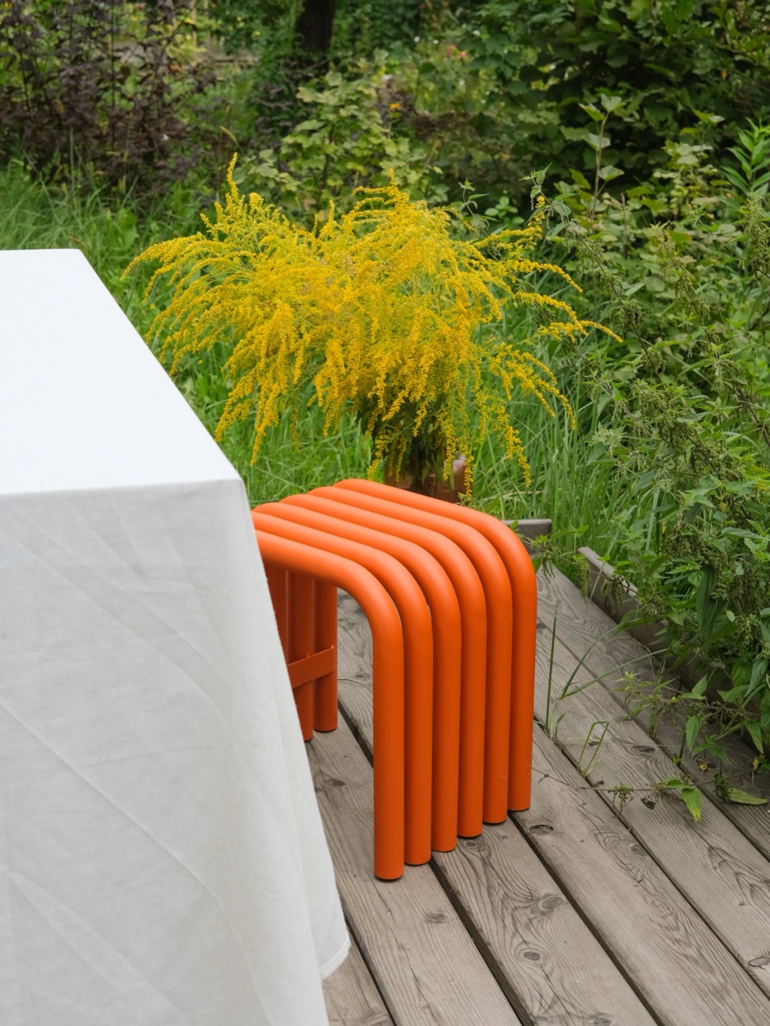 A minimalist Nokk Stool in a bright Orange Peel finish sits on a wooden deck. The stool has a bent-tube design and is next to a table covered with a white tablecloth. A plant with vibrant yellow flowers is visible behind the stool, with a lush green garden in the background.