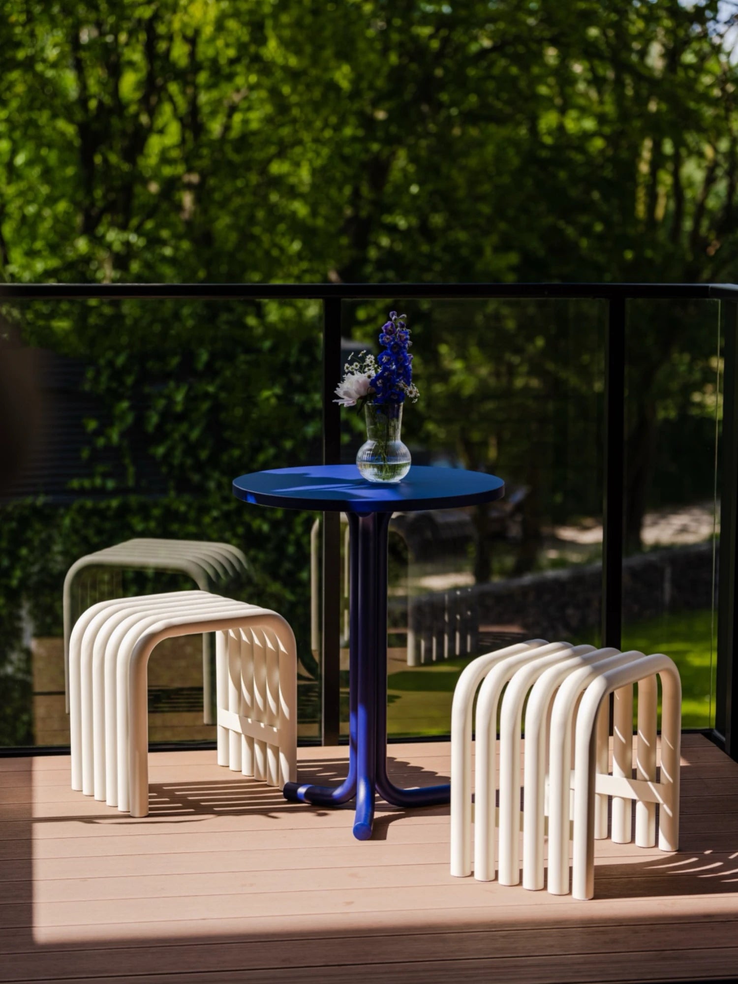A small outdoor furniture set is on a sunlit wooden deck. The set includes a dark blue pedestal table and two light Piazza Beige Nokk Stools, all made from bent tubes. A small glass vase with purple and white flowers is on the table, and lush green trees and a glass railing are visible in the background.