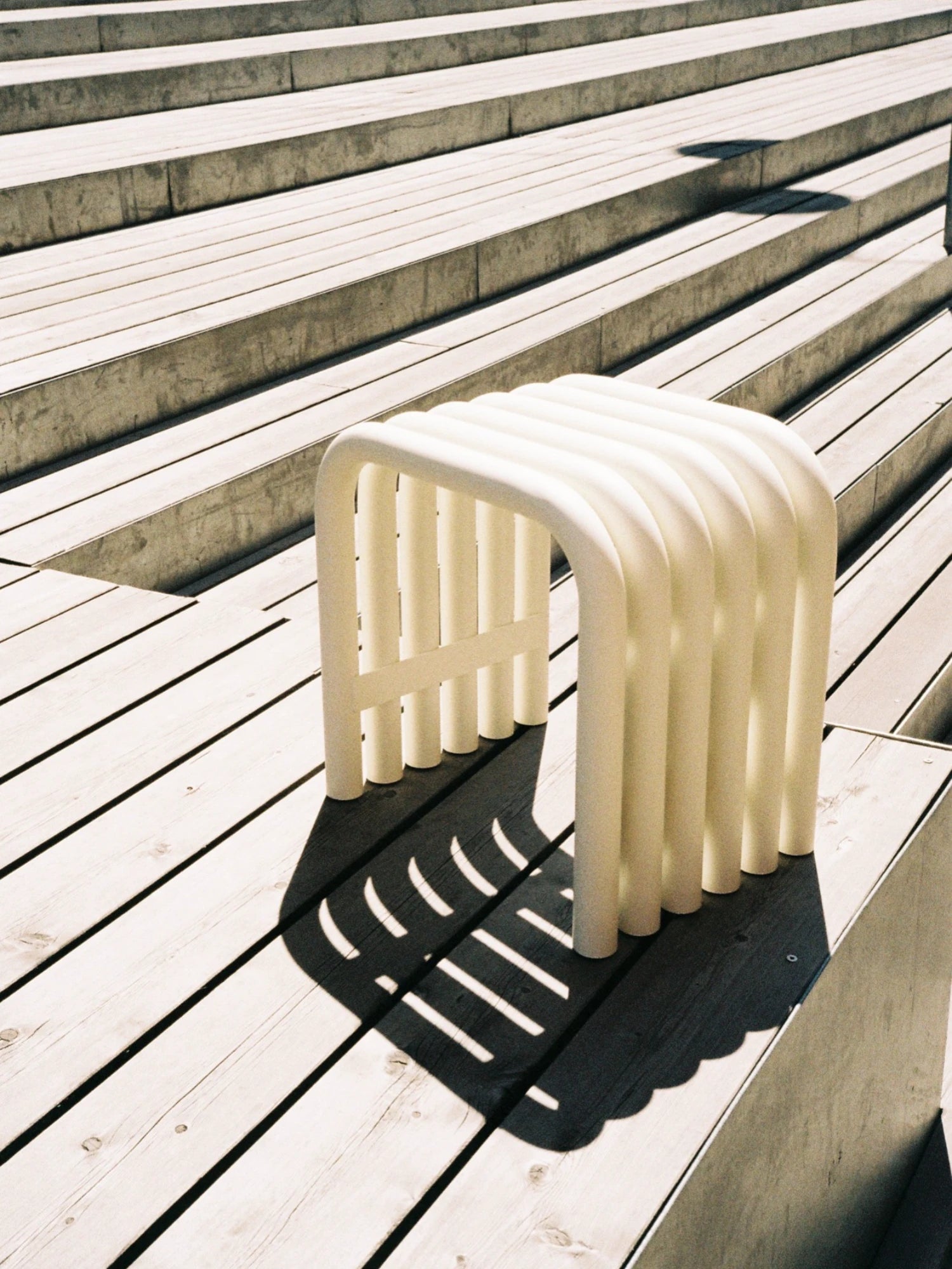 A light Piazza Beige Nokk Stool with a bent-tube design is placed on a wooden deck in the sunlight. The stool casts a striped shadow, and a set of concrete steps is visible in the background.