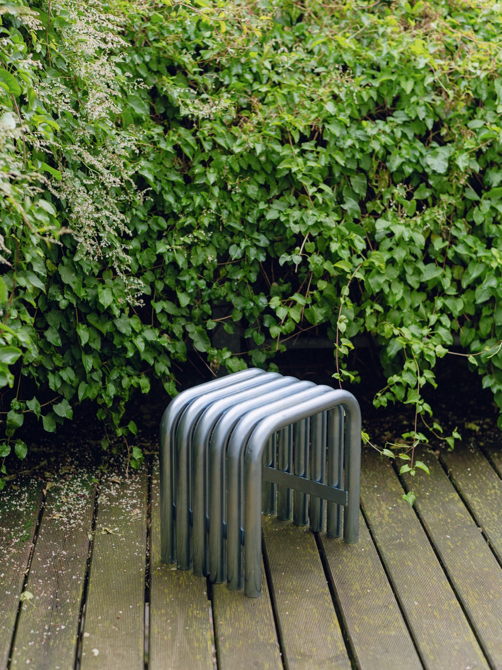 noo.ma Nokk Stool, an industrial chic metal stool crafted from multiple bent matte grey powder-coated steel tubes, creating a distinctive ribbed, minimalist design. This contemporary outdoor stool or accent table offers durable, modern seating. Ideal for garden patios, balconies, or as brutalist-inspired indoor furniture.